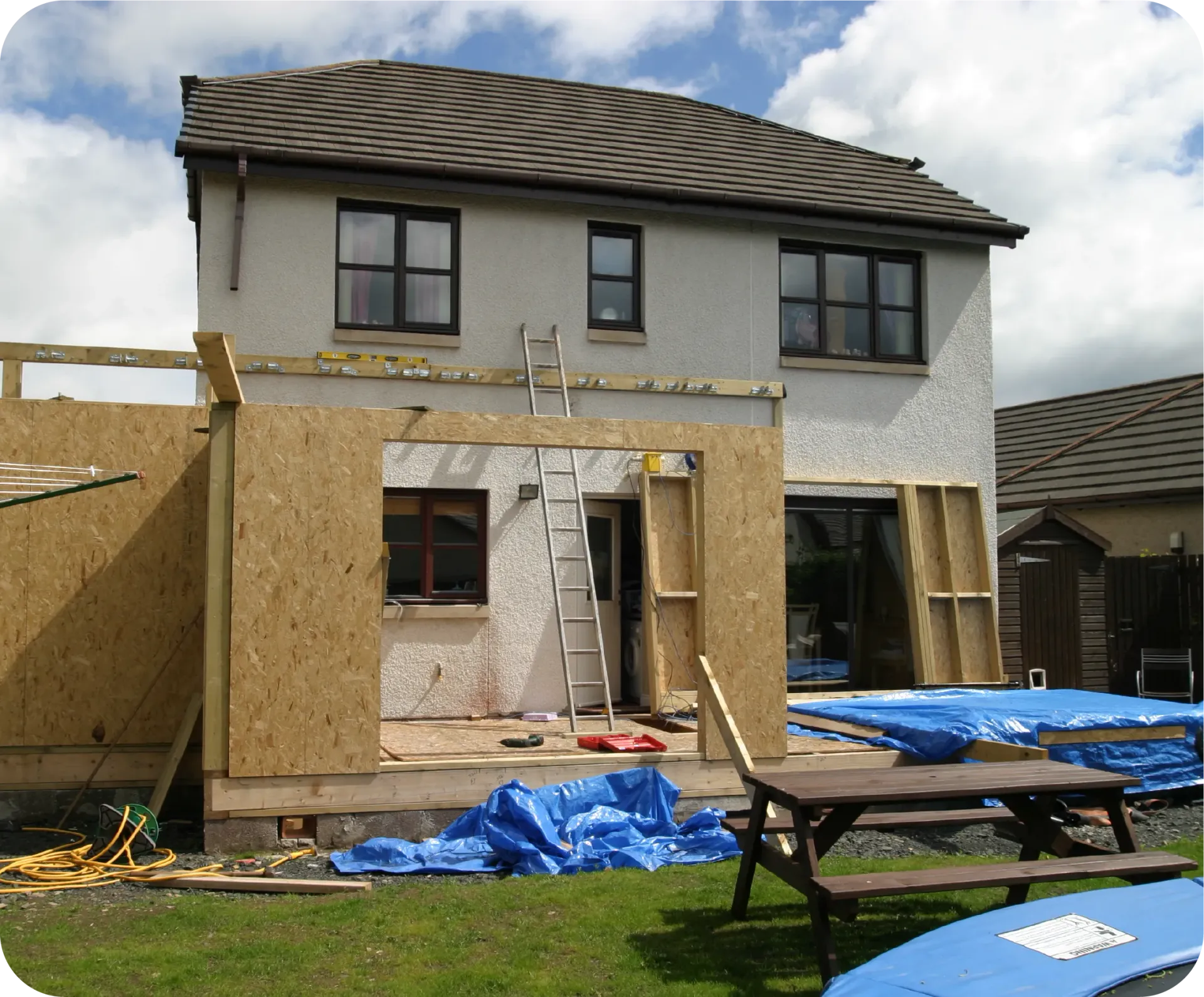 House under construction with a wooden frame extension, blue tarps, and outdoor setting.
