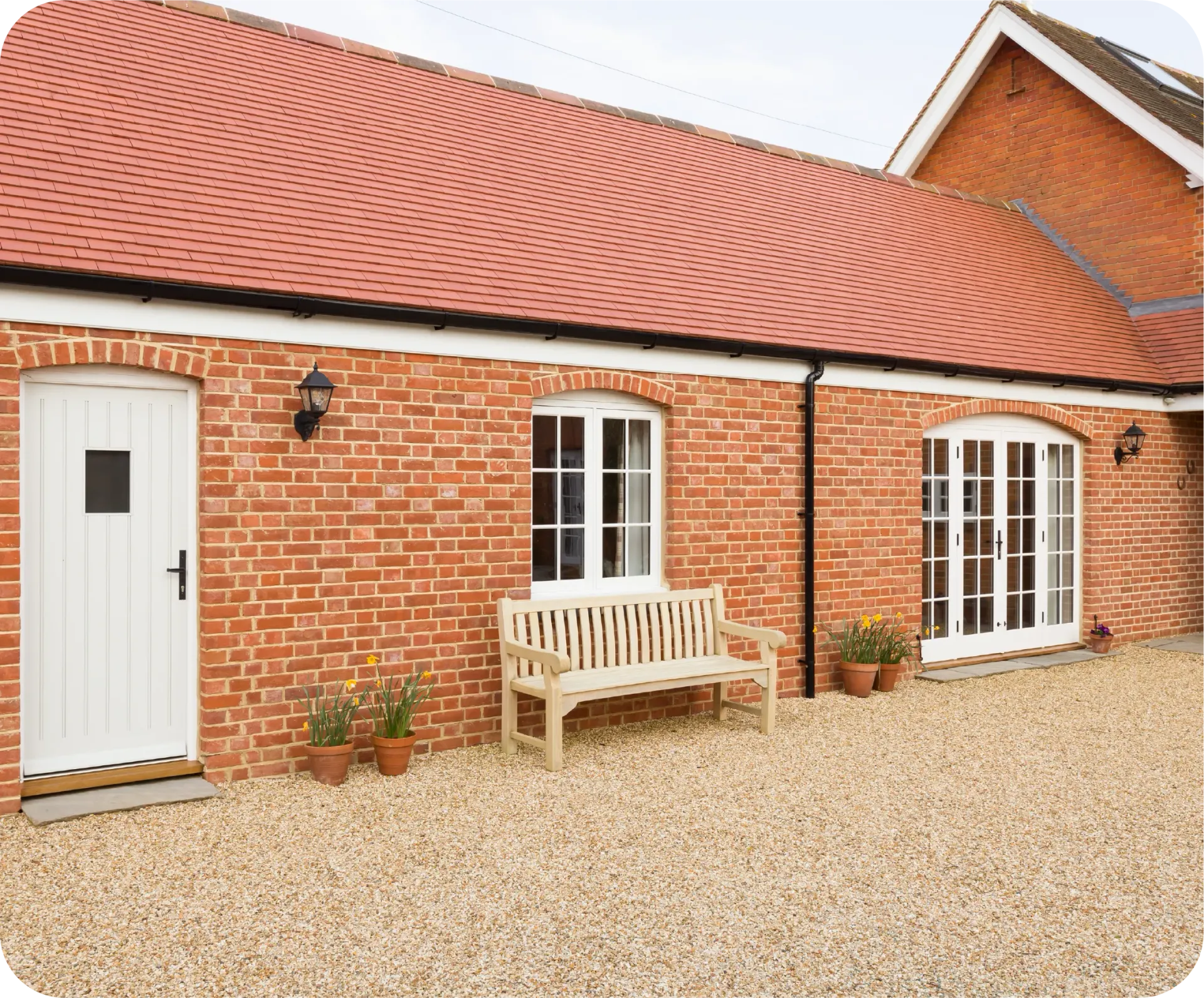 Brick building with red tile roof, white door and windows, wooden bench, and gravel yard.