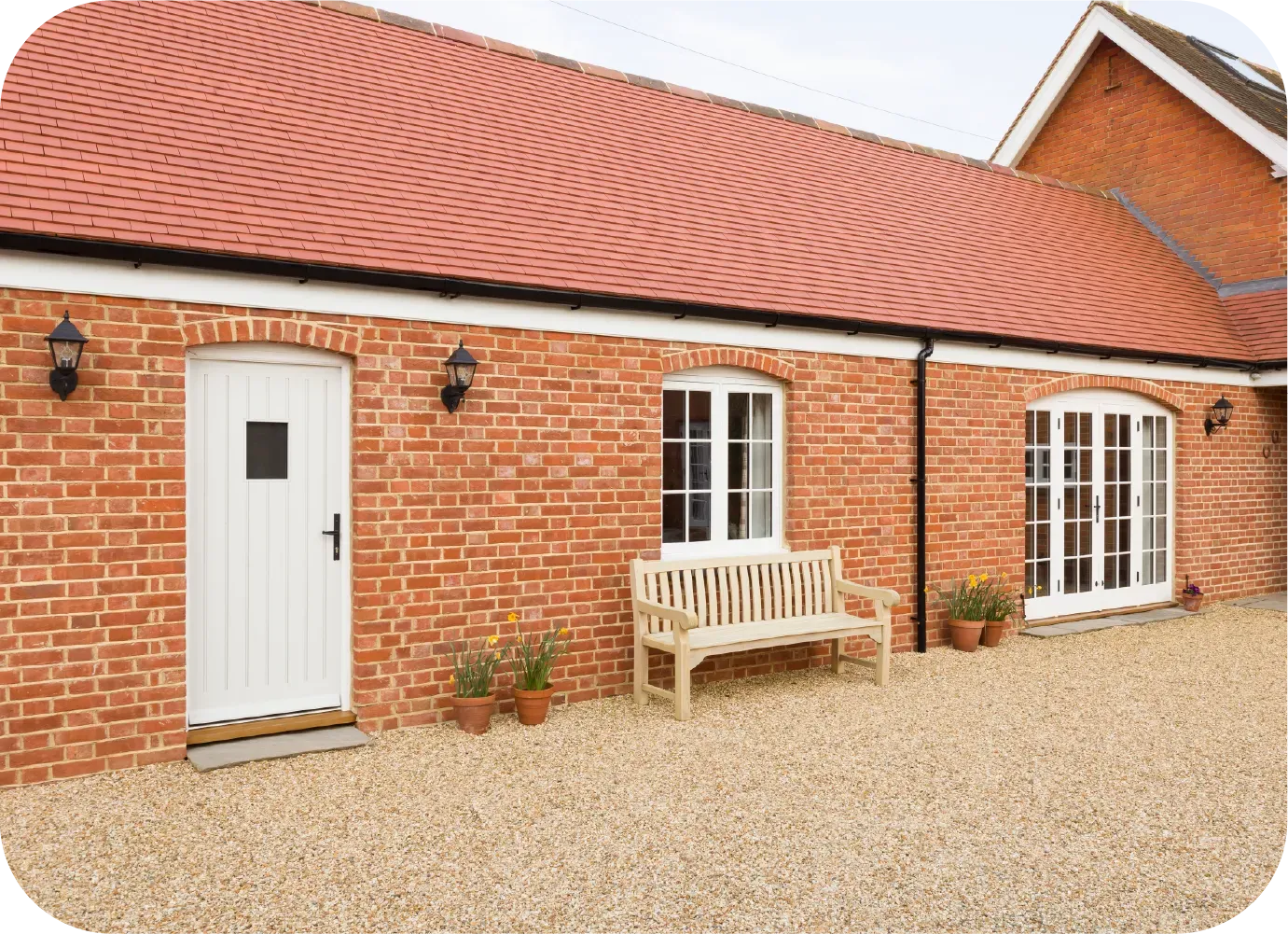 Brick building with red tile roof, white door, window, and bench on a gravel patio.