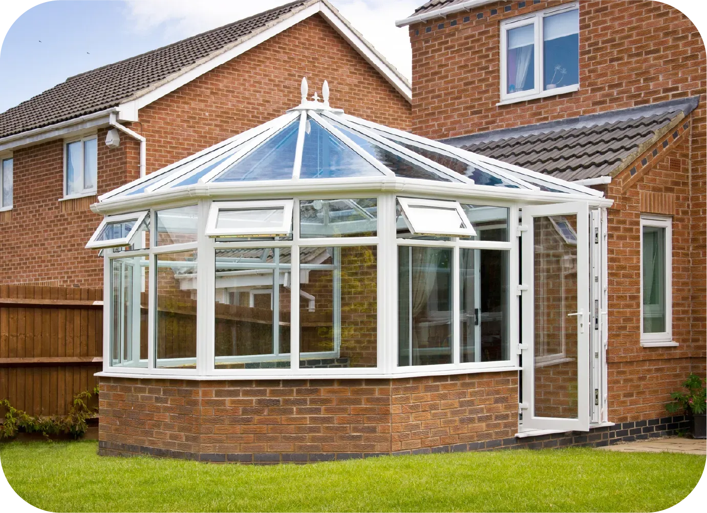 White-framed glass conservatory with brick base, attached to a brick house, on green lawn.