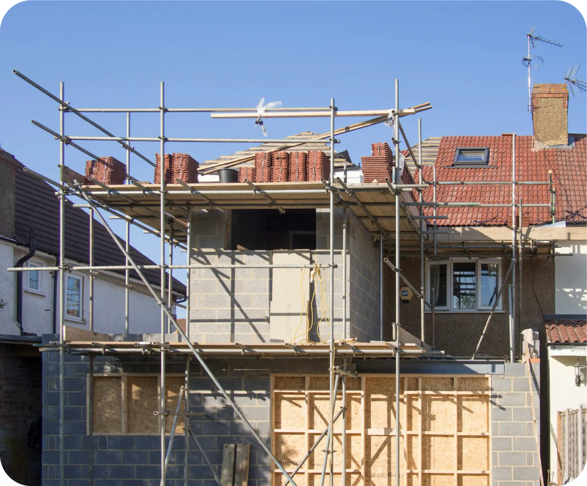 Scaffolding surrounds a building under construction, with bricks on the platform and a blue sky overhead.