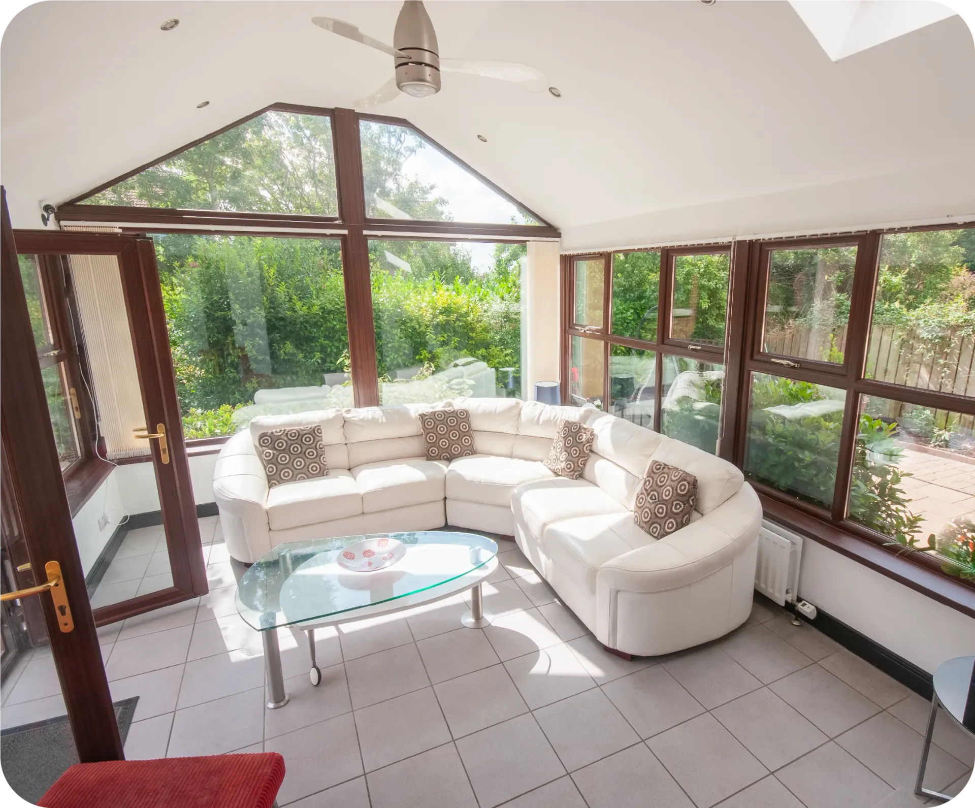 Sunroom with white sectional sofa, glass coffee table, and large windows overlooking greenery.