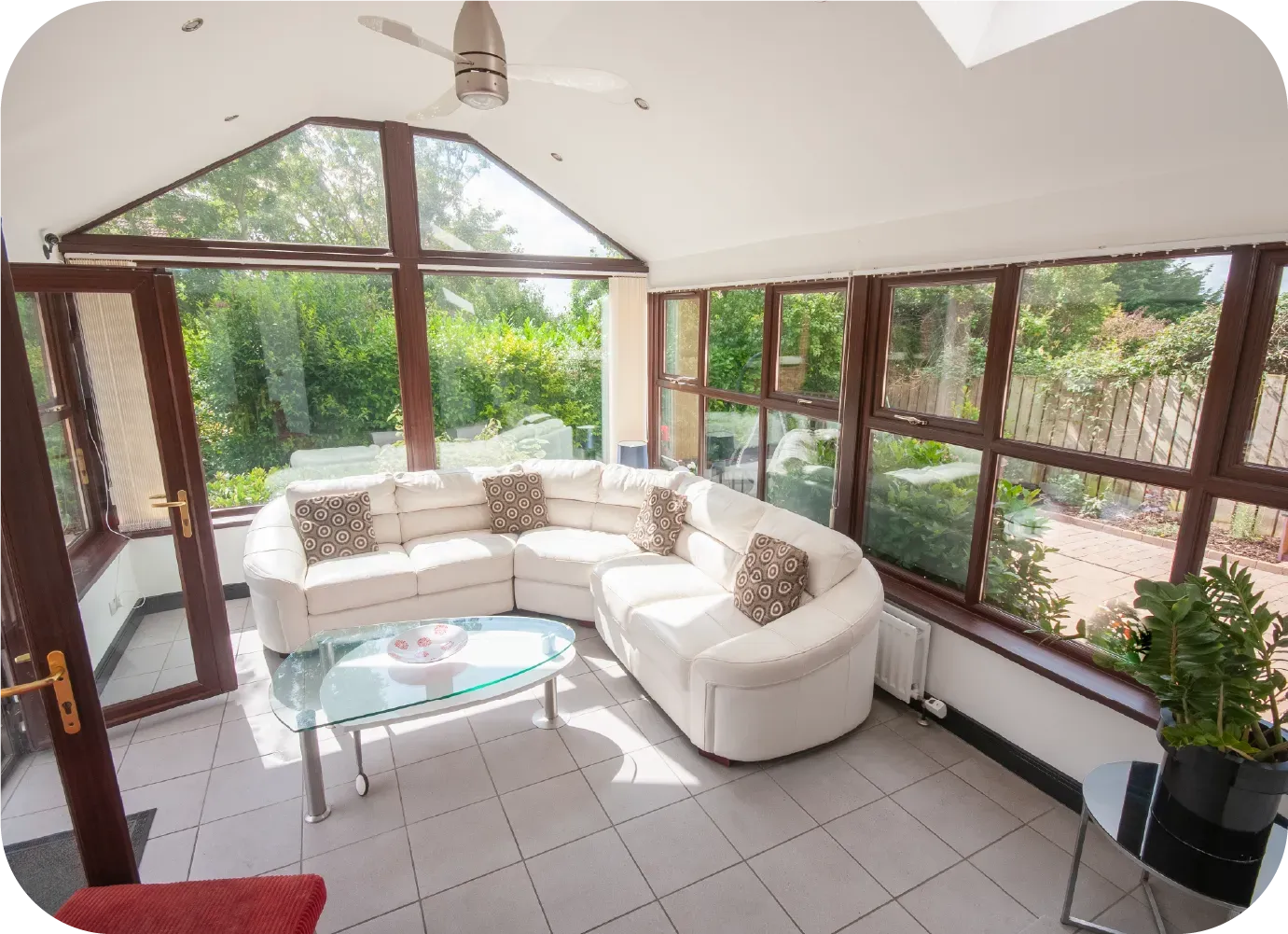 Sunroom with white sectional sofa, glass table, and tiled floor, surrounded by windows.