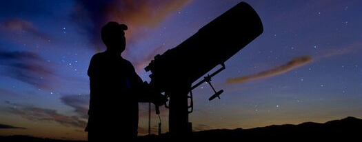 A silhouette of a person observing the night sky through a large telescope at twilight.