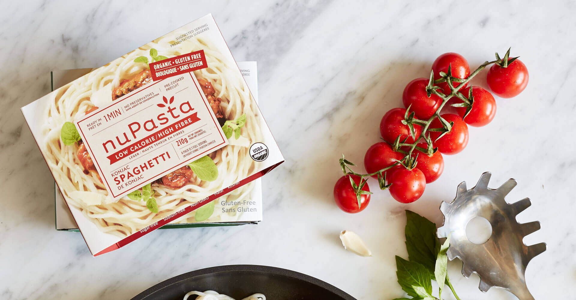 A box of nupasta spaghetti is sitting on a table next to a pan of spaghetti and tomatoes.