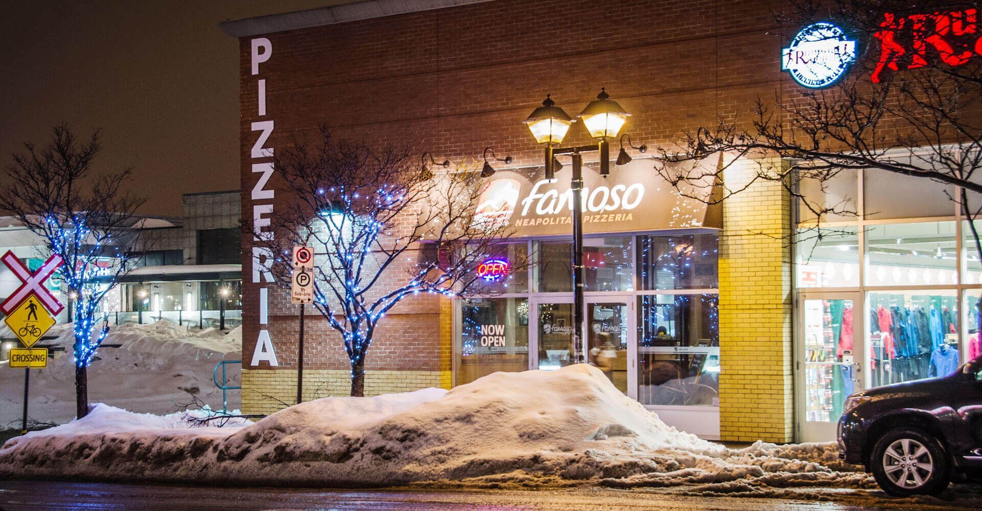 A car is parked in front of a pizzeria in the snow.