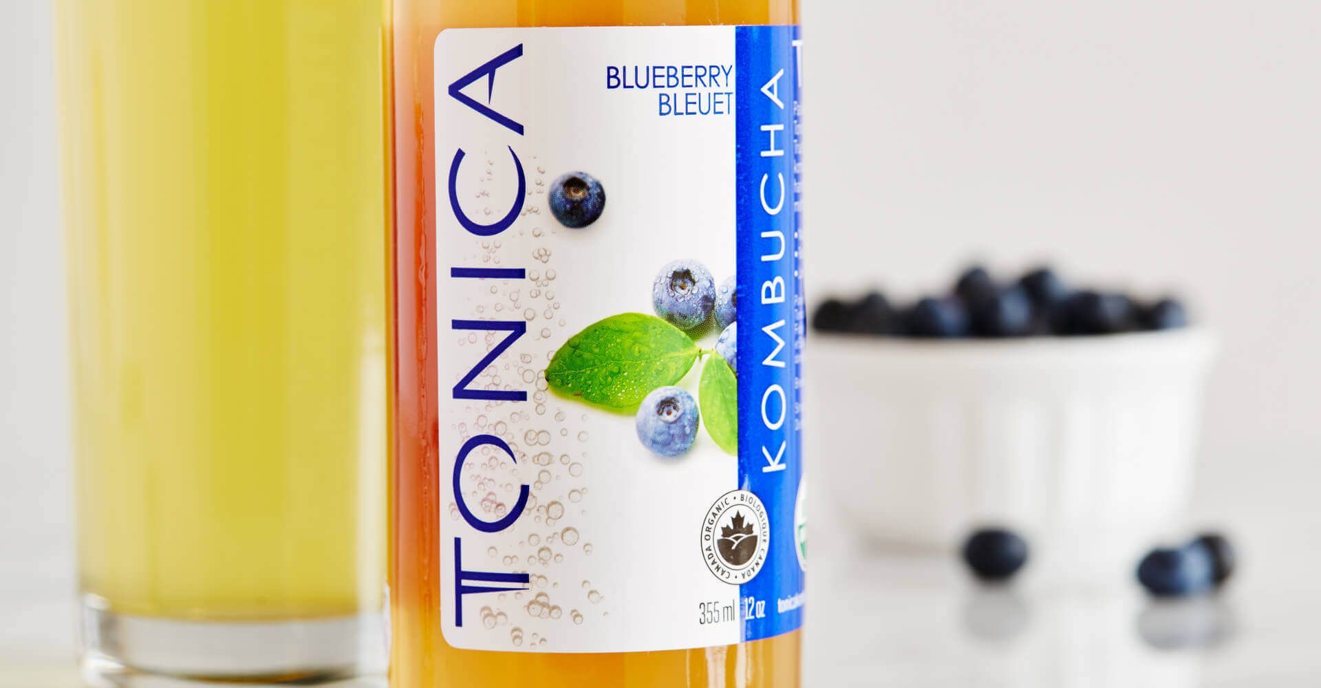 A bottle of kombucha next to a cup of blueberries.