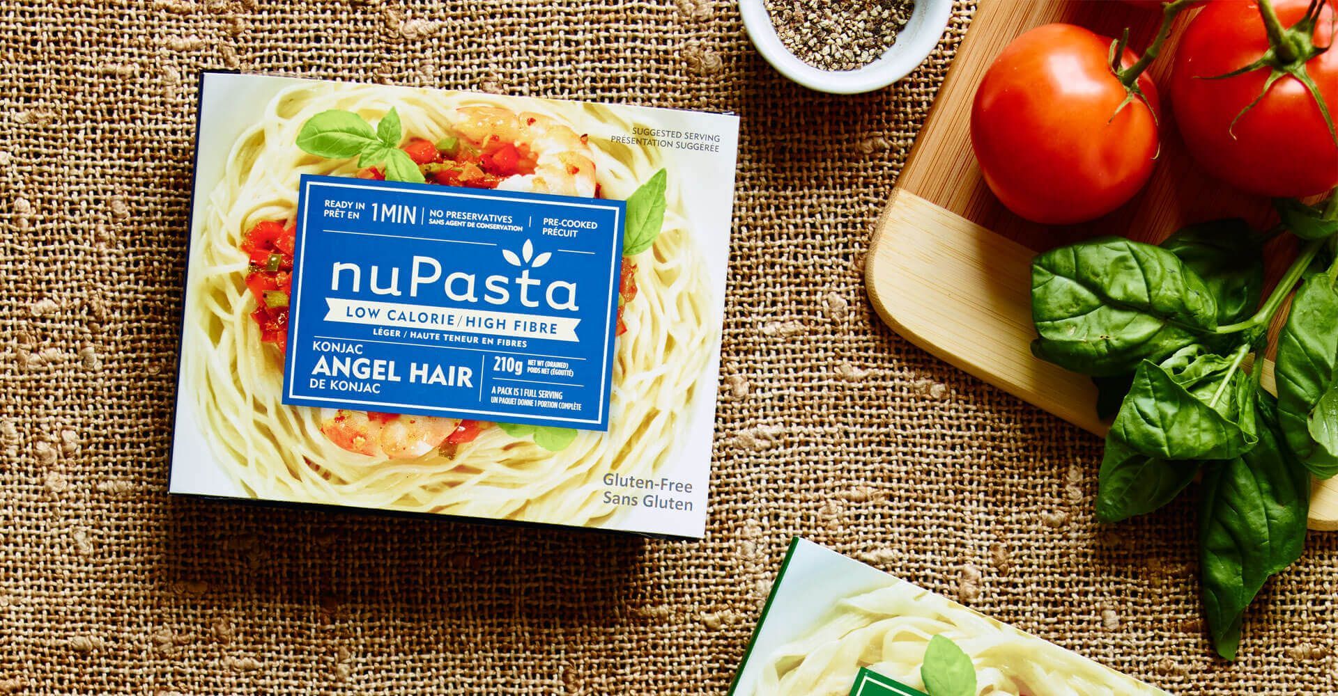 A box of nupasta spaghetti is sitting on a table next to tomatoes and spinach.