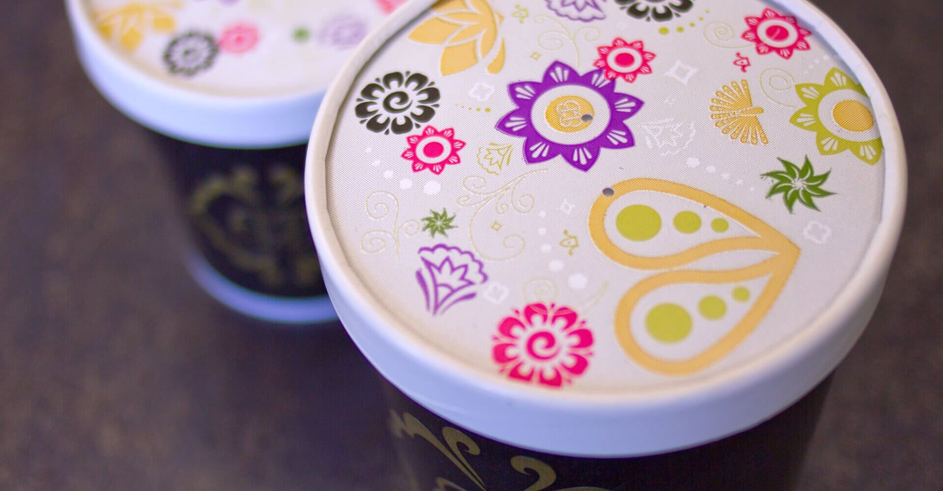 Two cups of coffee with flowers on the lids are sitting on a table.