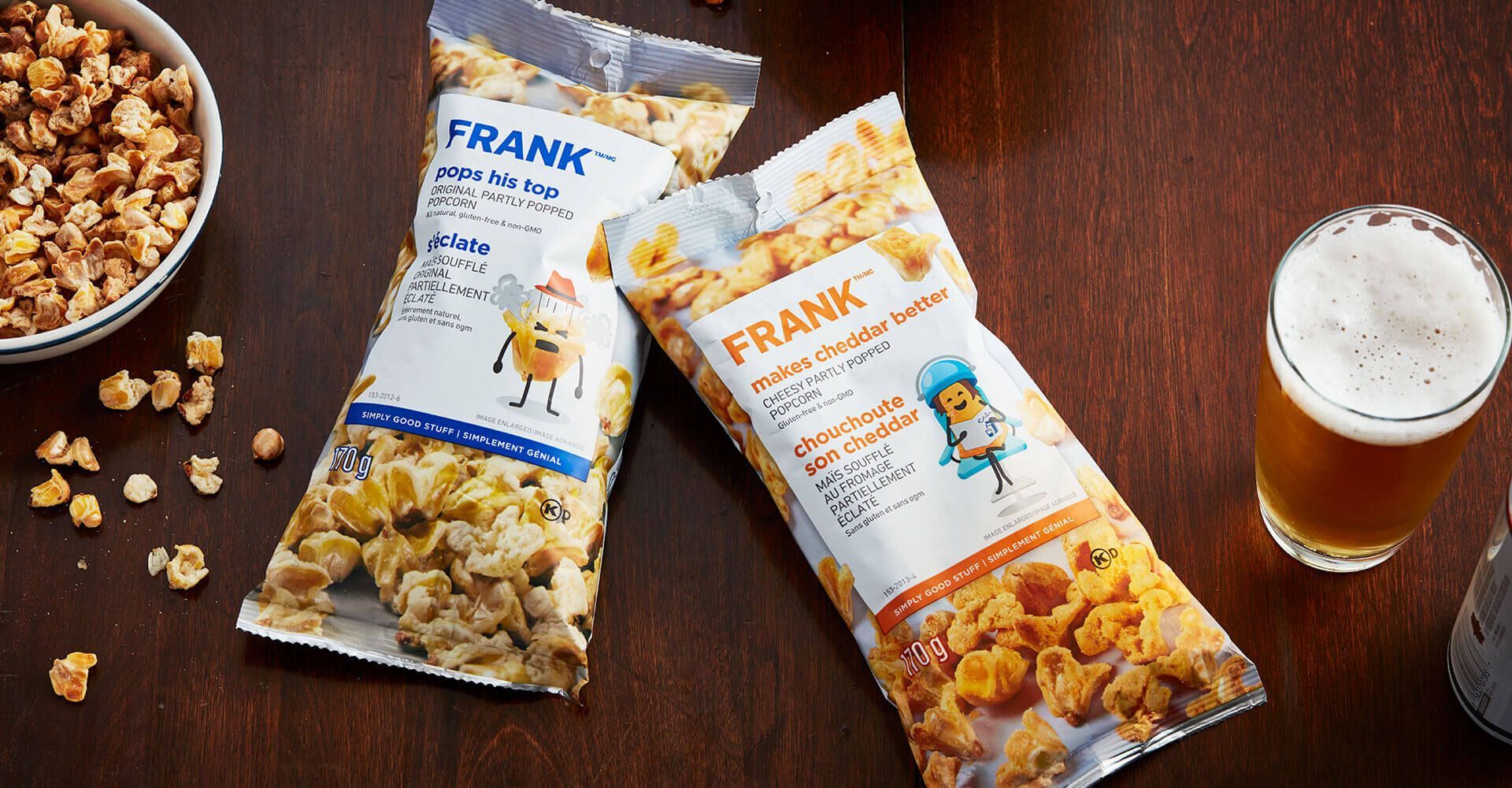 Two bags of frank 's caramel popcorn are sitting on a wooden table next to a glass of beer.