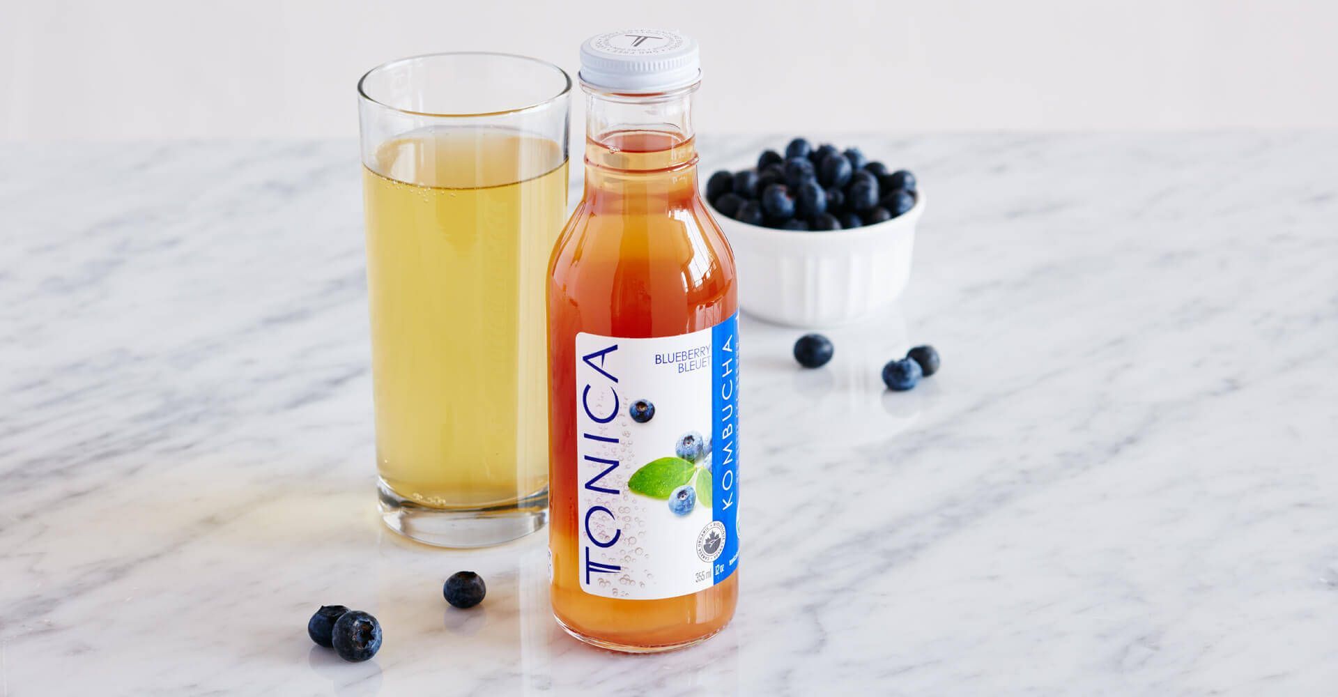 A bottle of tonica next to a glass of tonica and a bowl of blueberries.
