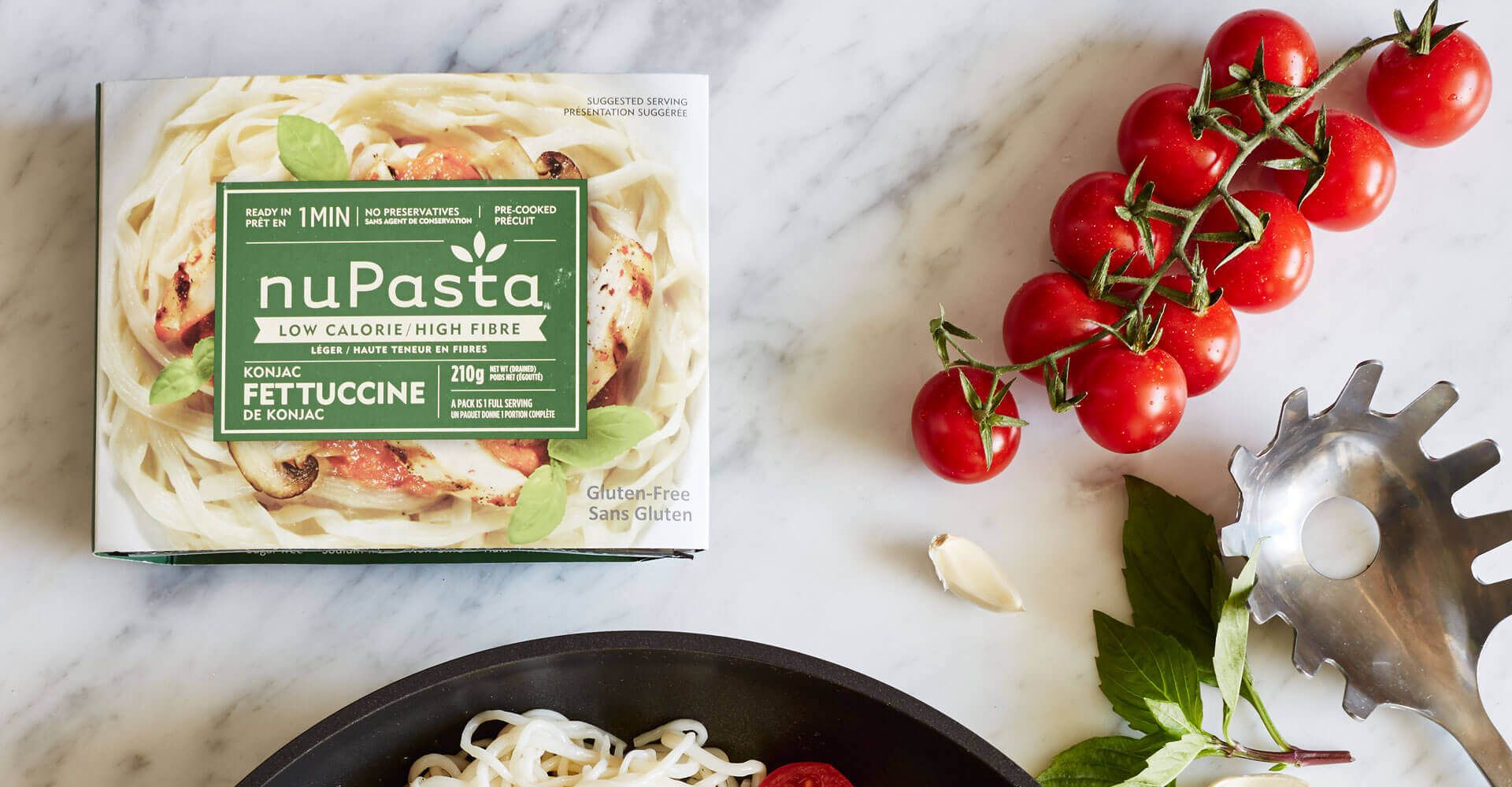 A box of nupasta pasta next to a pan of pasta and tomatoes.