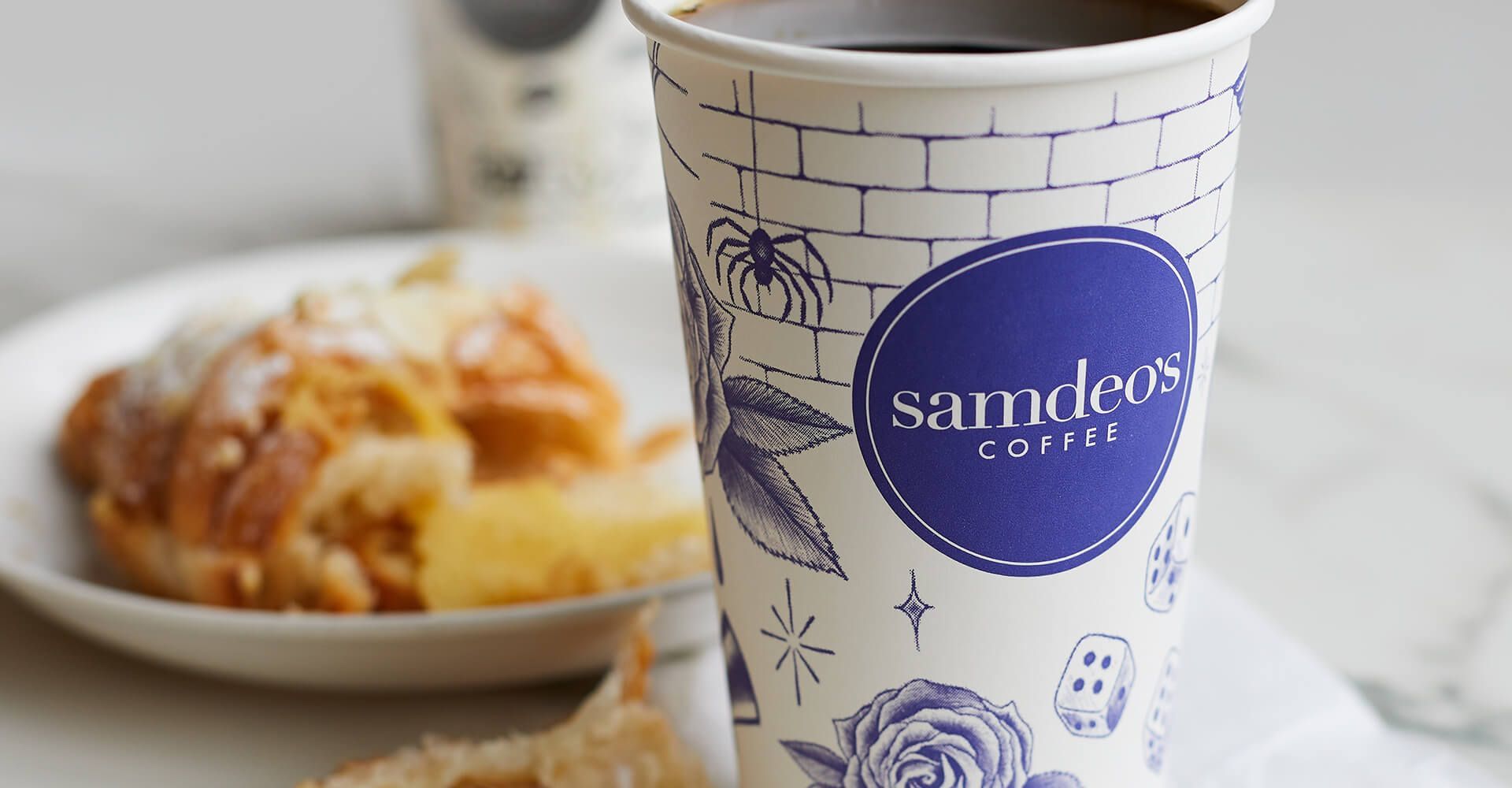 A cup of samdeos coffee is sitting on a table next to a plate of food.