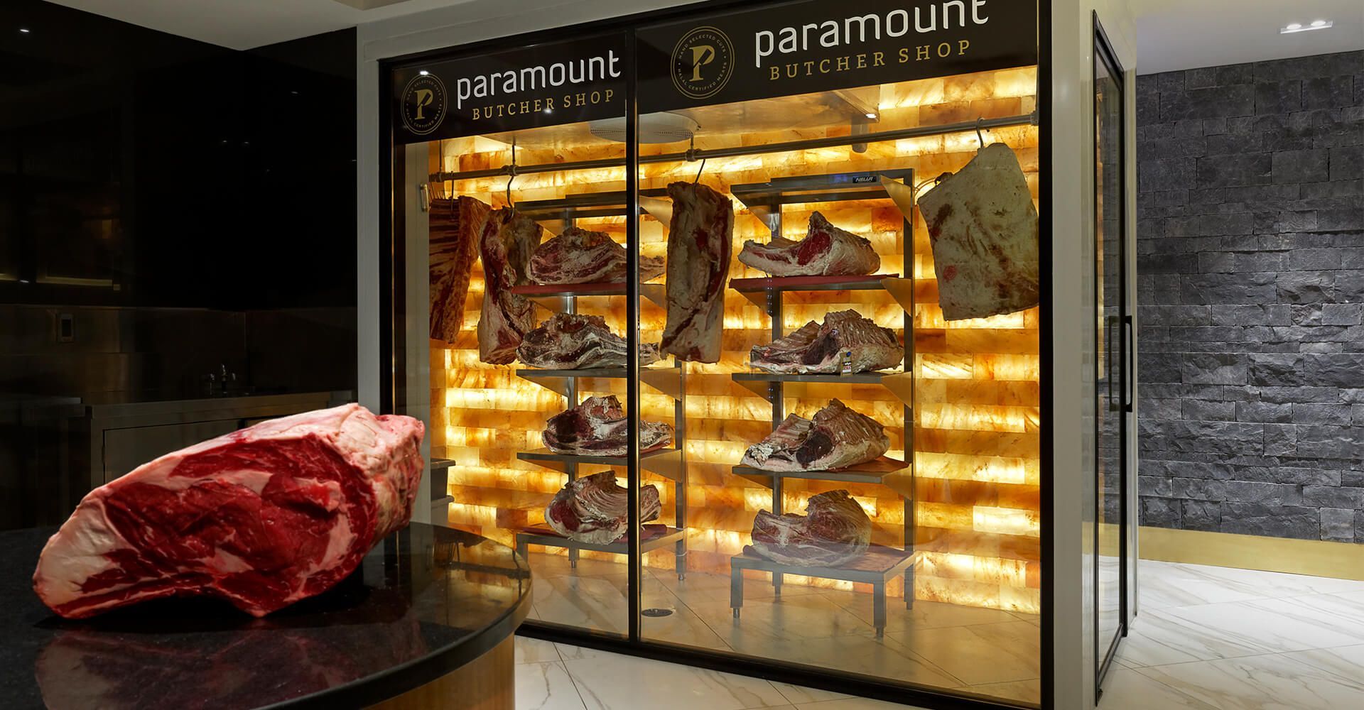A large piece of meat is sitting on a table in front of a refrigerator filled with meat.