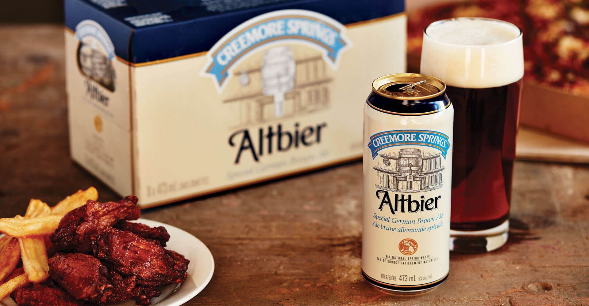 A can of altbier beer next to a box of chicken wings and a glass of beer.