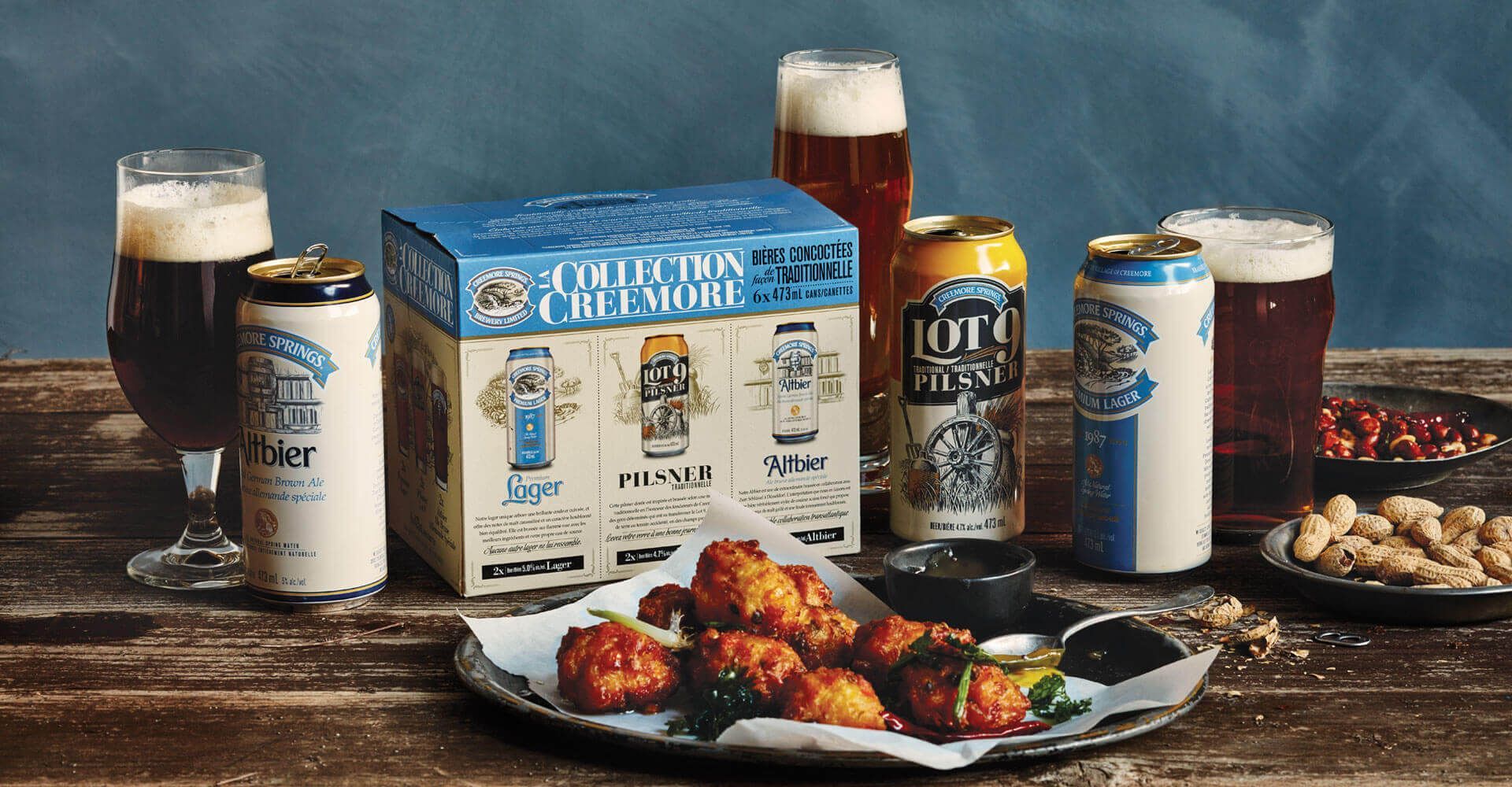 A box of beer is sitting on a wooden table next to a plate of fried chicken.