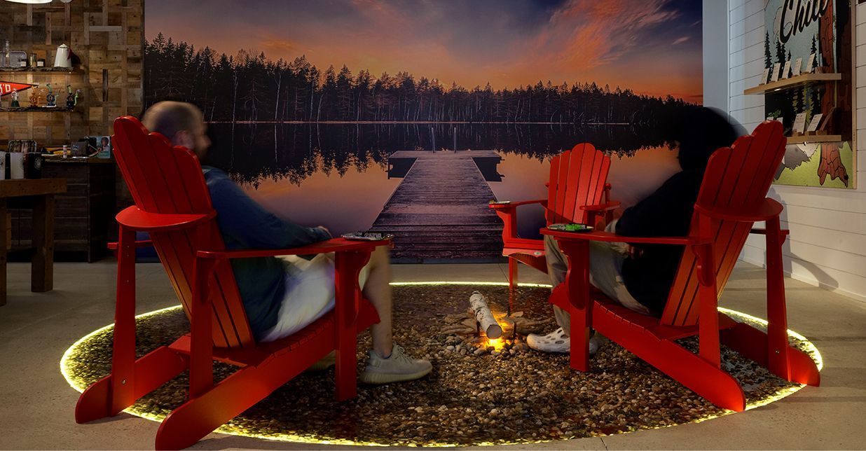 Two people are sitting in red adirondack chairs in front of a lake.