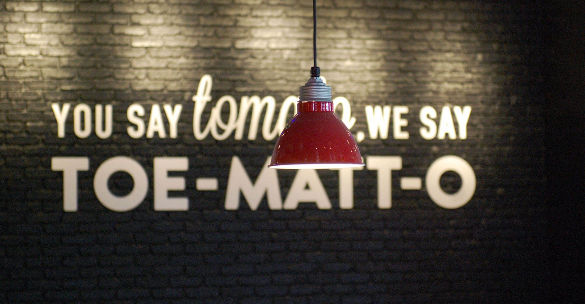 A sign on a brick wall says you say tomo we say toe-matt-o
