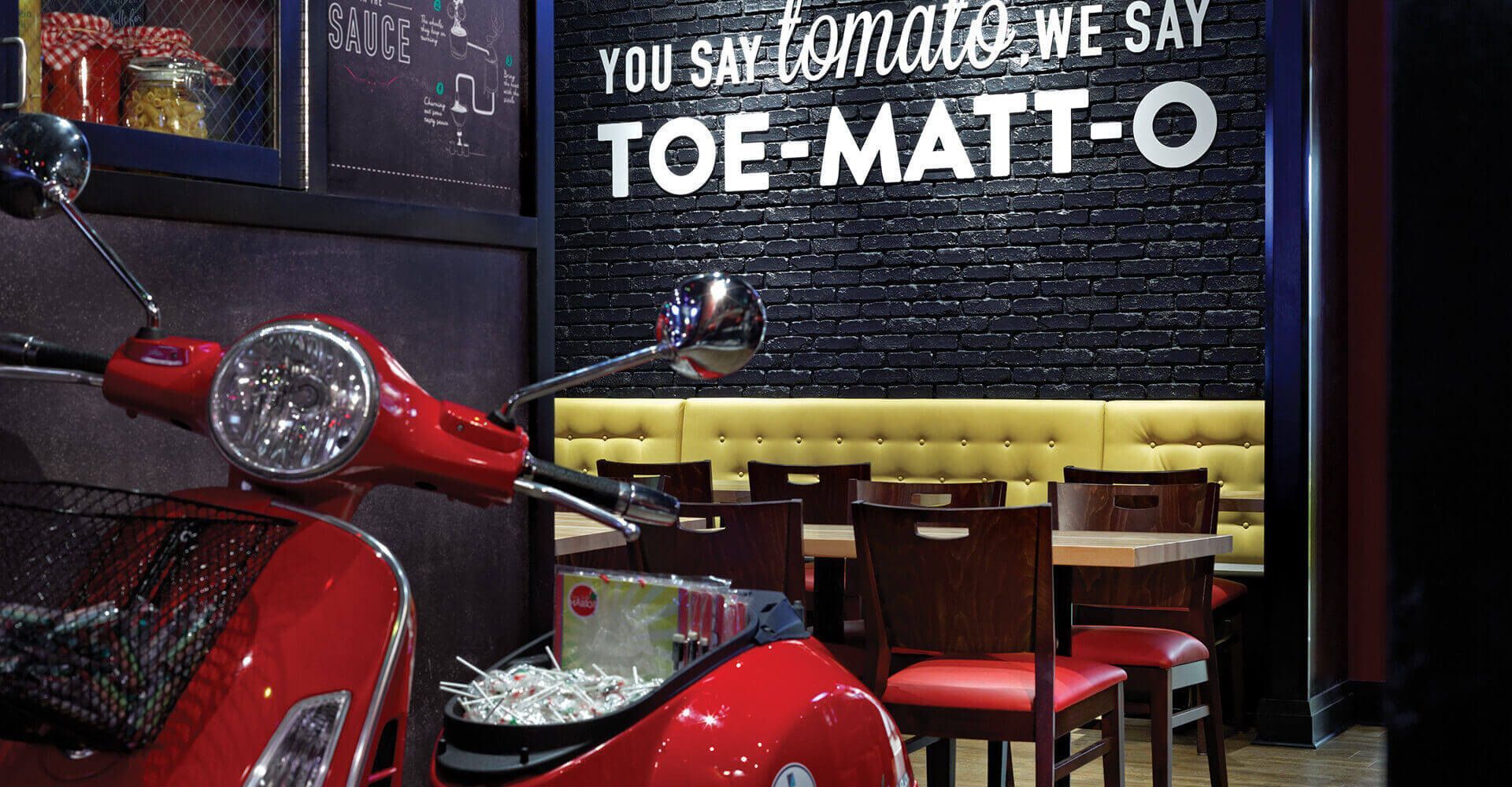 A red scooter is parked in front of a sign that says toe-matt-o