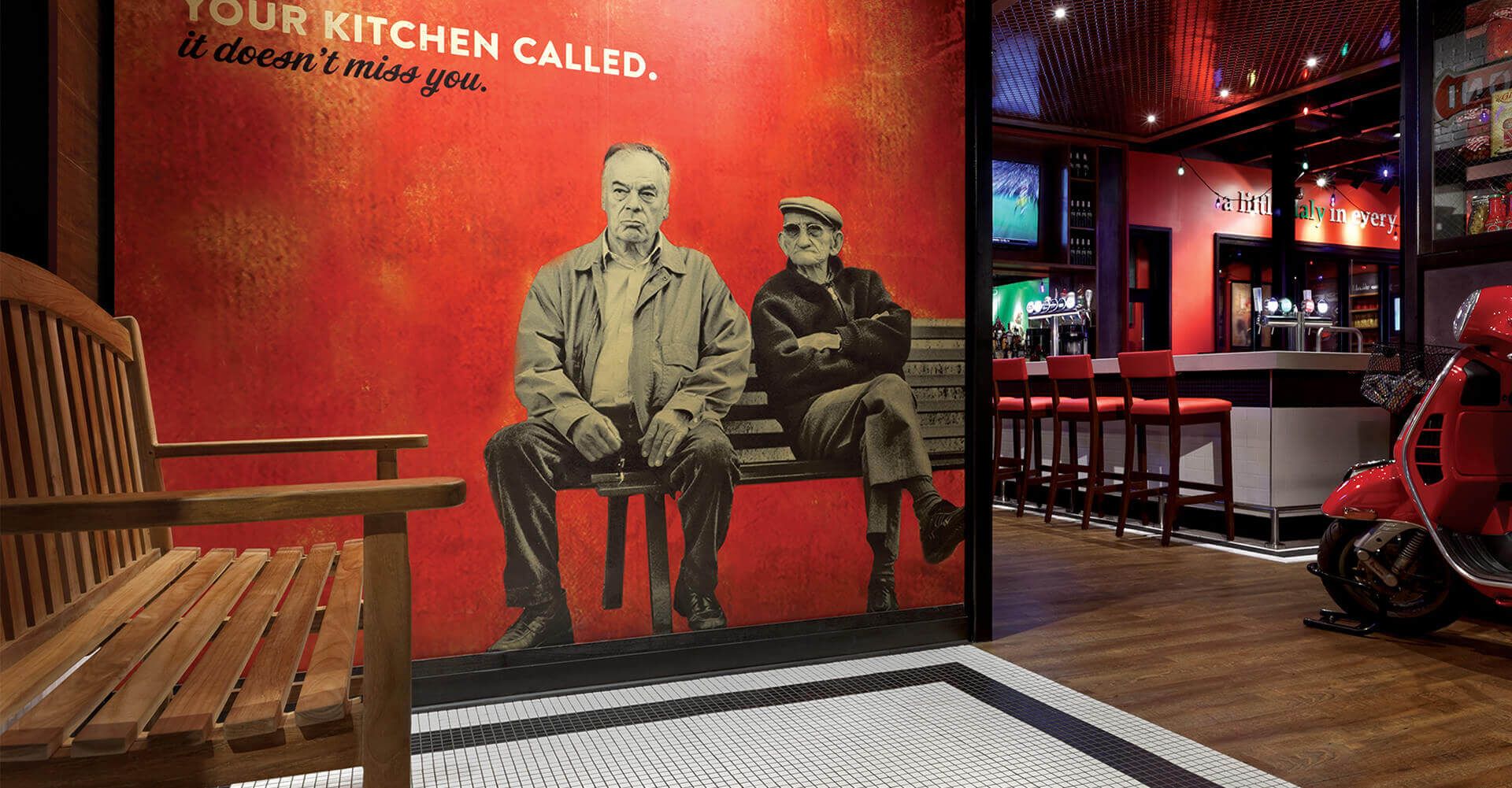Two men are sitting on a bench in front of a red wall.