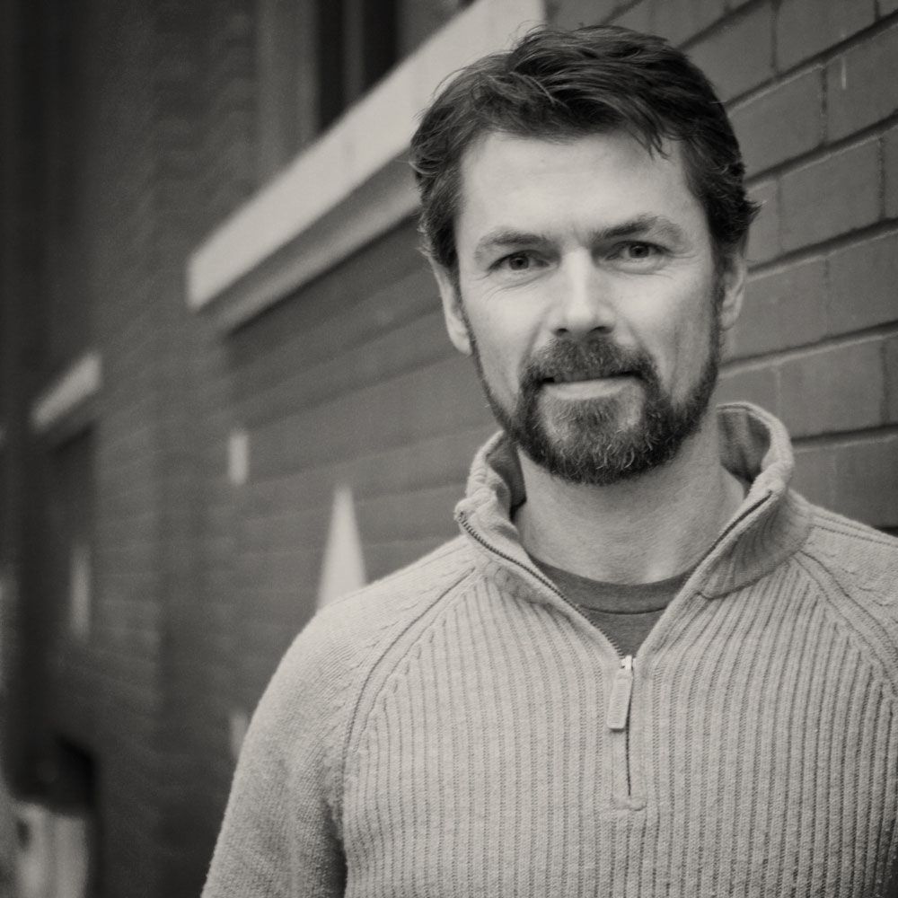 A man with a beard wearing a sweater is standing in front of a brick wall.
