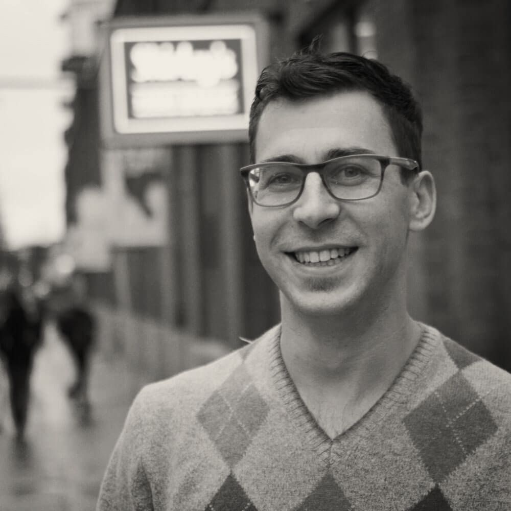 A man wearing glasses and a plaid sweater is smiling in a black and white photo.