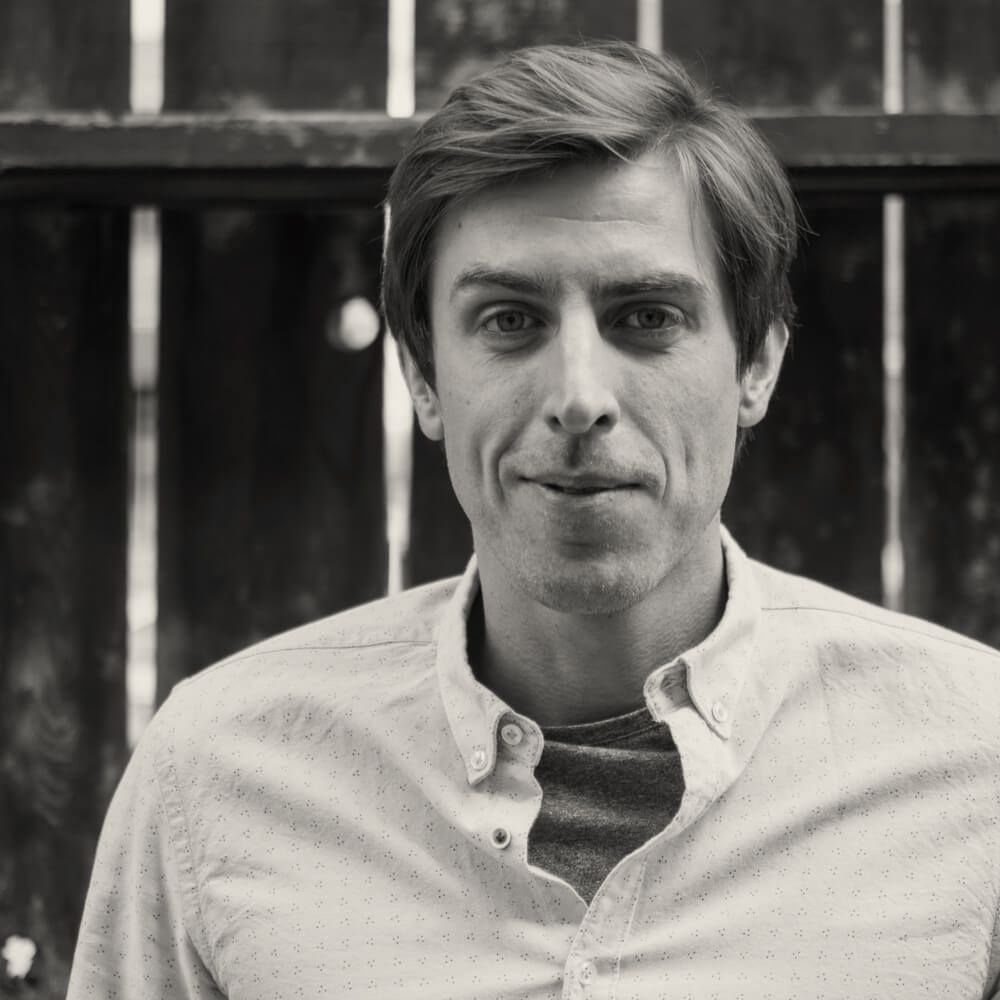 A man in a white shirt is standing in front of a wooden fence.