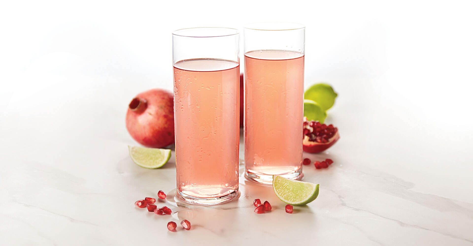 Two glasses of pomegranate juice with limes and pomegranate seeds on a table.