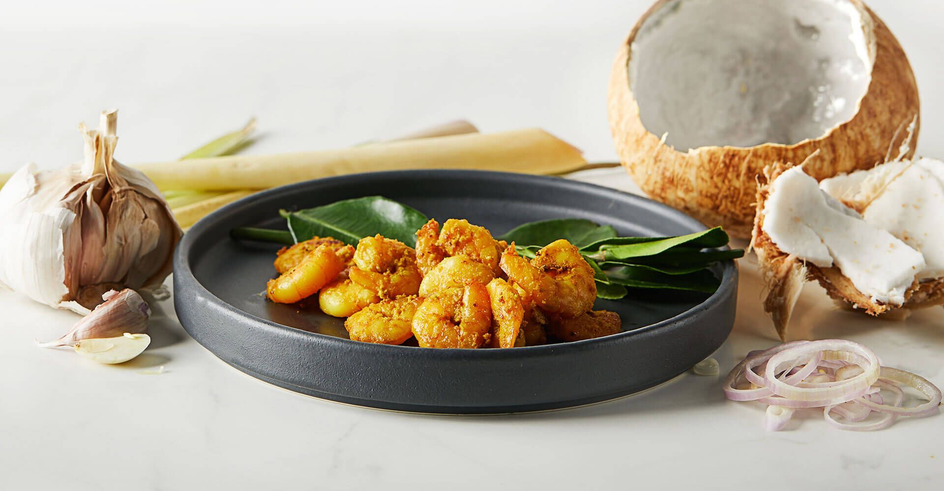 A plate of shrimp is sitting on a table next to a coconut and garlic.