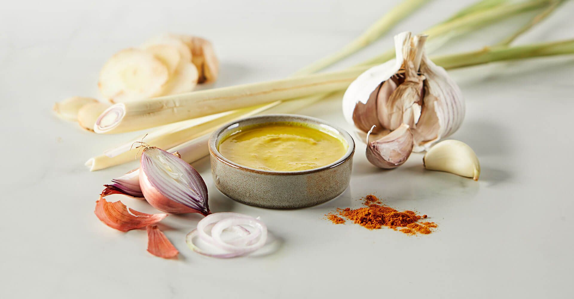 A bowl of sauce surrounded by garlic , lemongrass , onions , and red pepper.