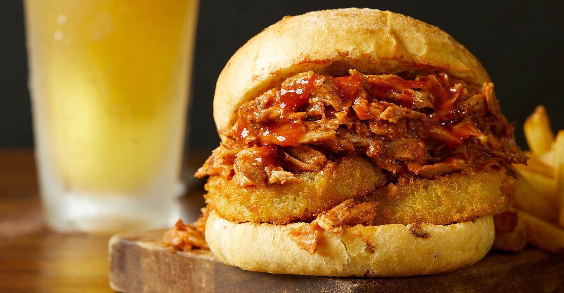 A pulled pork sandwich is sitting on a wooden cutting board next to a glass of beer.