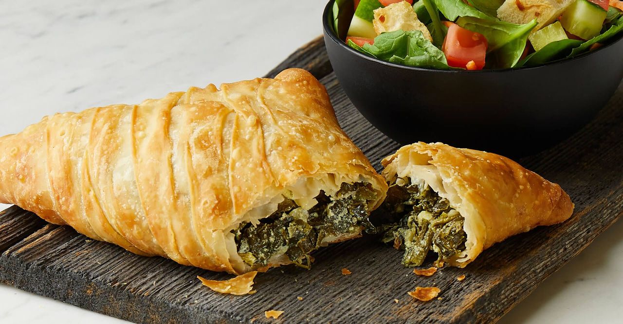 A pastry with spinach and cheese is sitting on a wooden cutting board next to a bowl of salad.