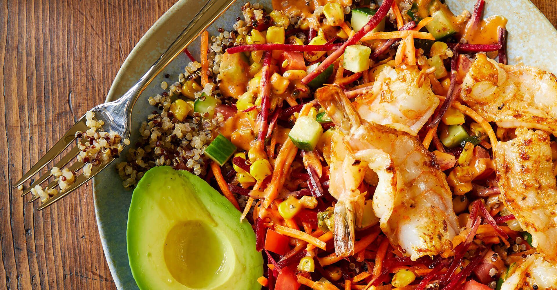 A plate of food with shrimp , avocado , rice and vegetables on a wooden table.