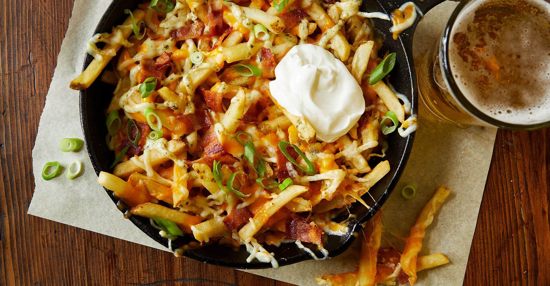 A bowl of french fries with sour cream and bacon next to a glass of beer.