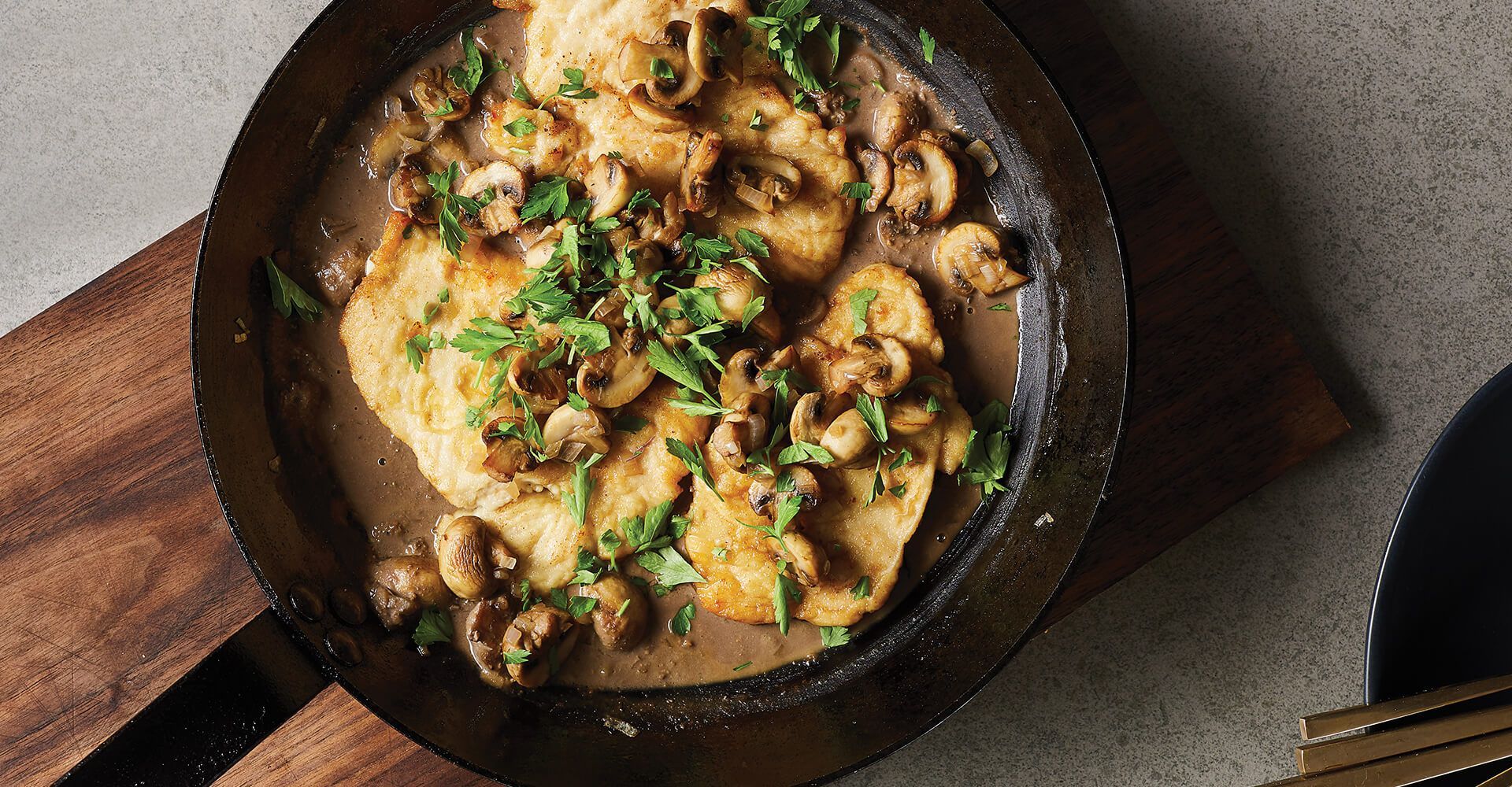 A pan filled with chicken and mushrooms on a wooden cutting board.
