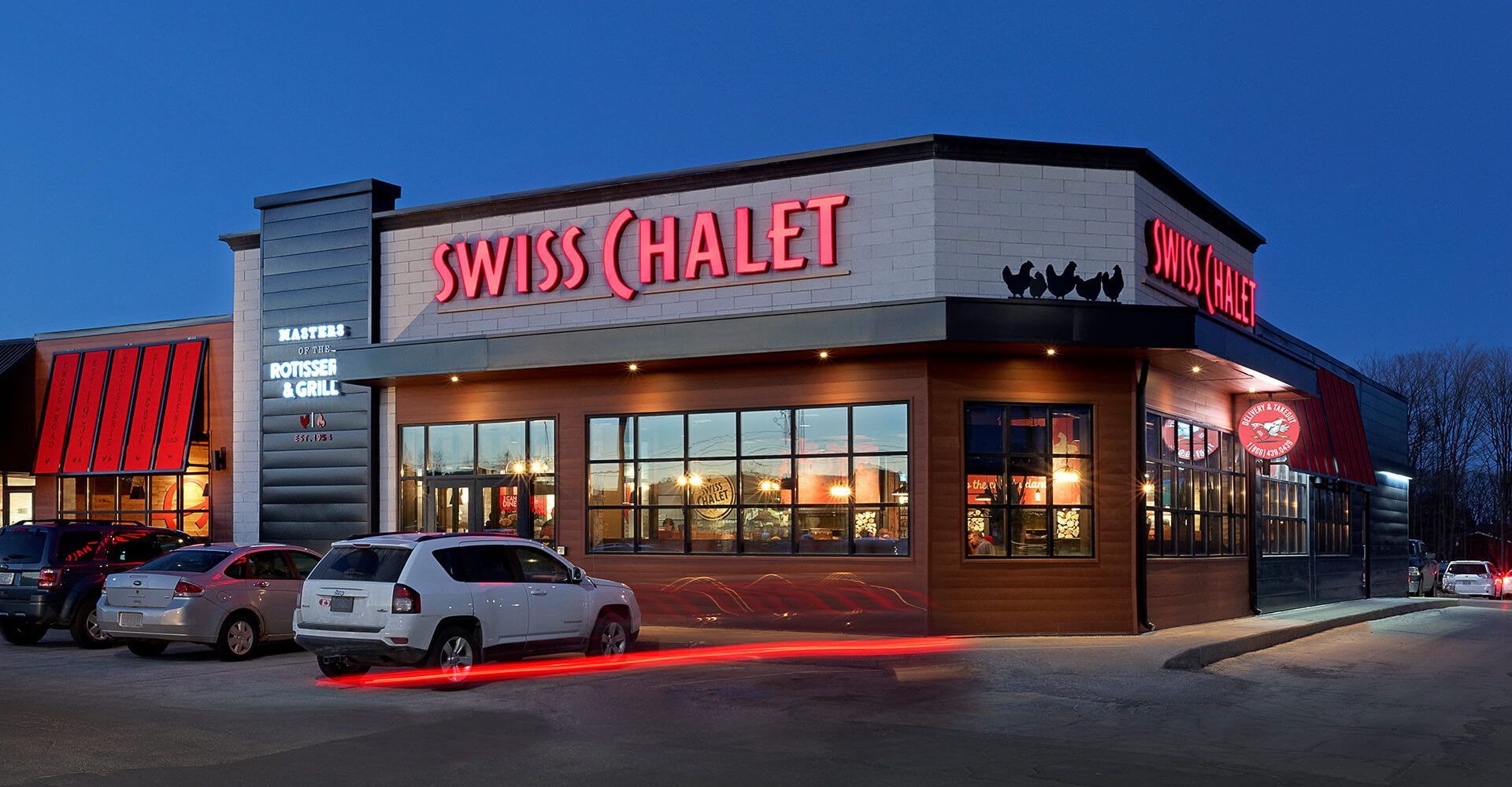 A swiss chalet restaurant with cars parked in front of it at night.