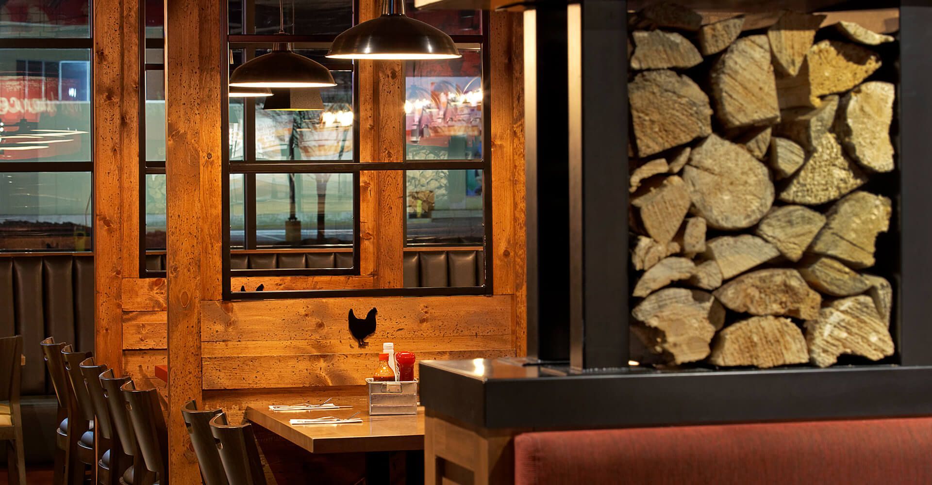 A restaurant with wooden tables and chairs and a pile of logs on the wall.