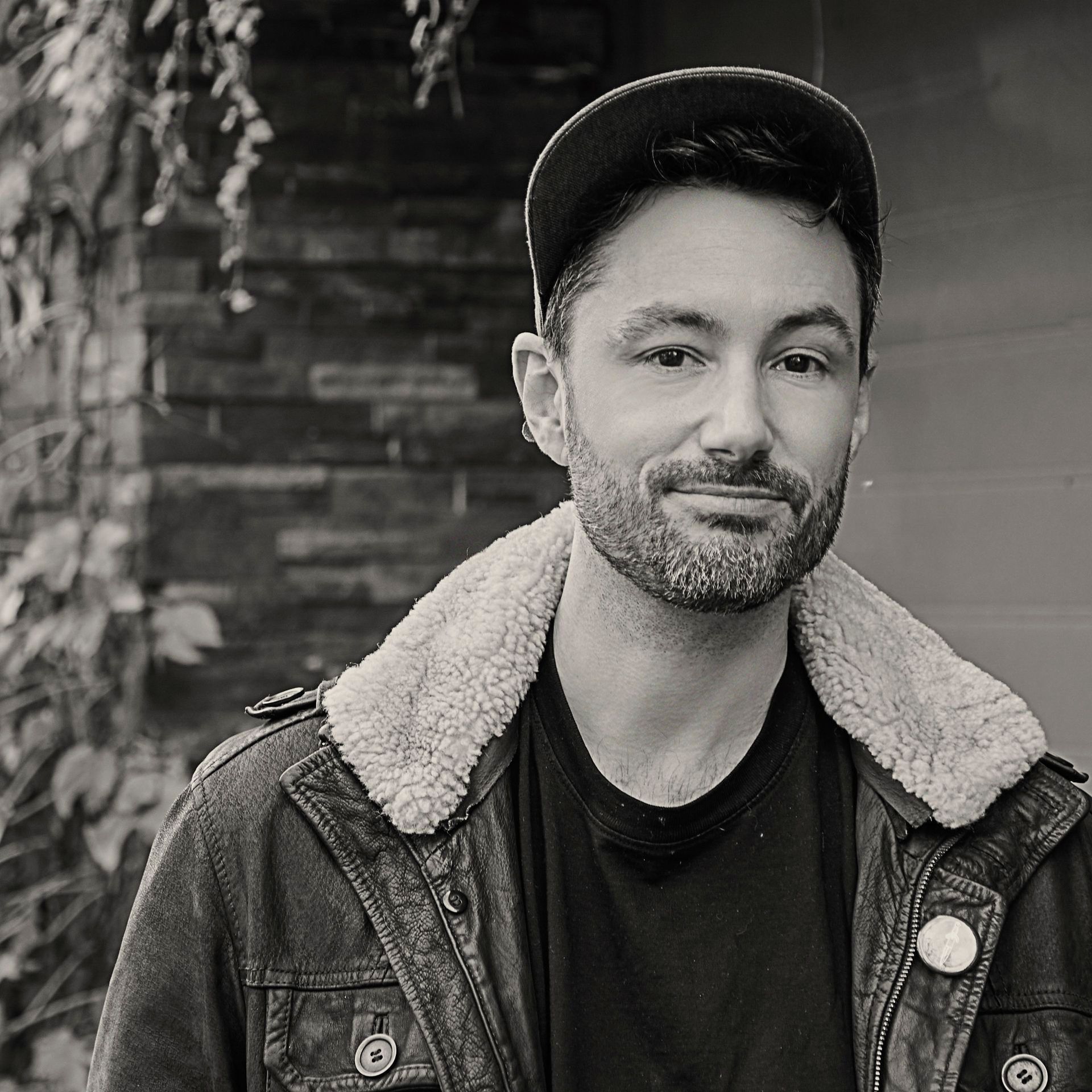 A man with a beard is wearing a hat and a leather jacket.
