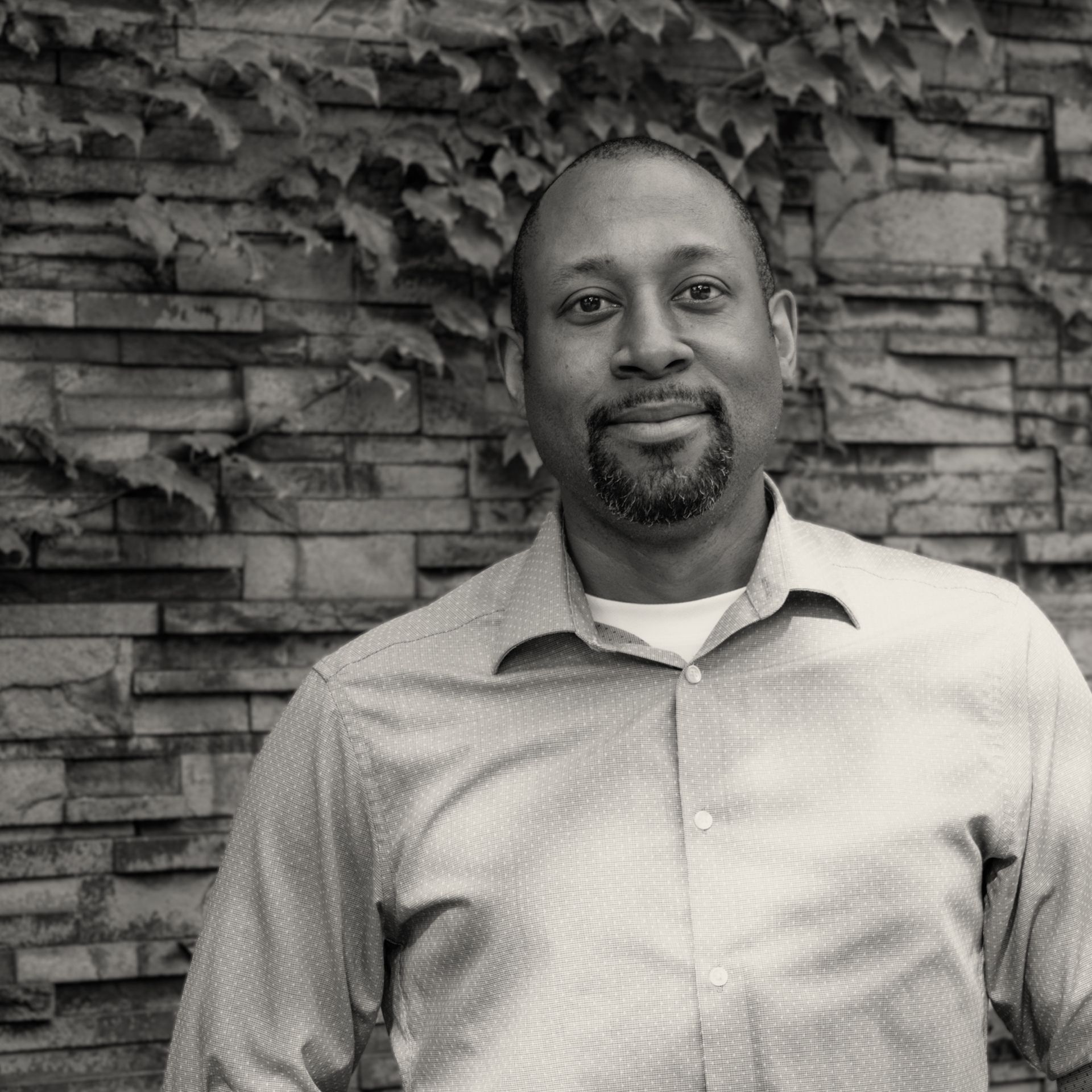 A man with a beard is standing in front of a brick wall in a black and white photo.