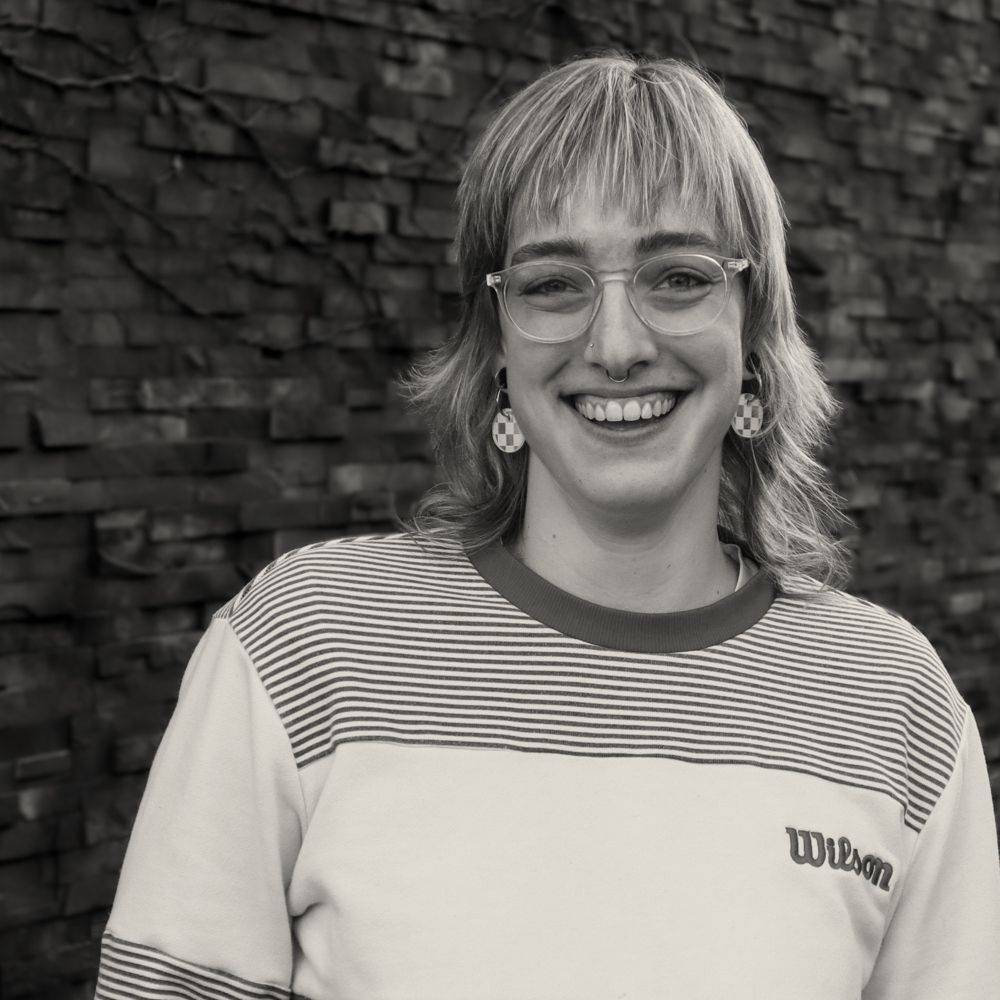 A woman wearing glasses and a wilson shirt is smiling in front of a brick wall.