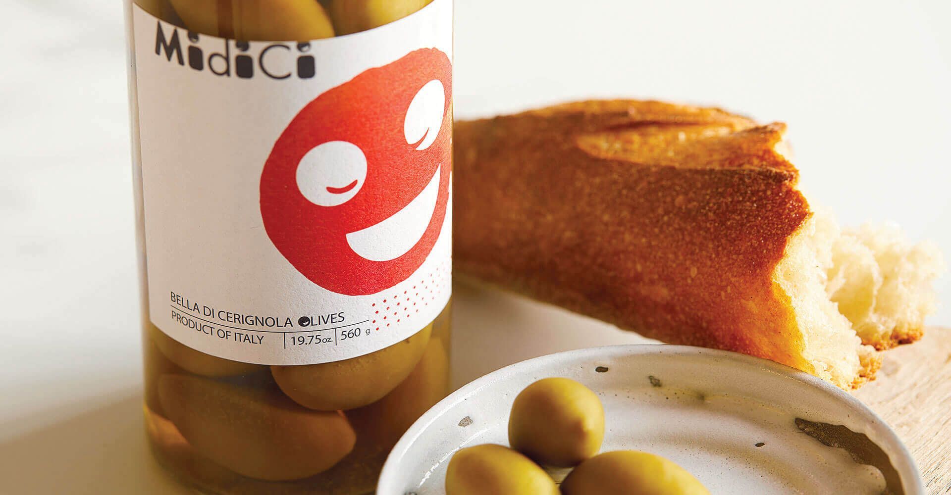 A jar of olives next to a plate of olives and a piece of bread.