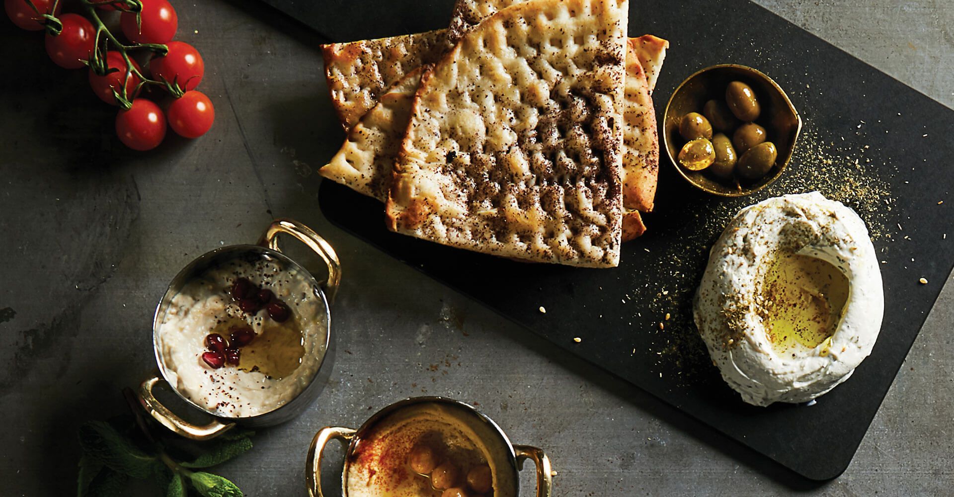 A cutting board topped with bread , hummus , olives and tomatoes.