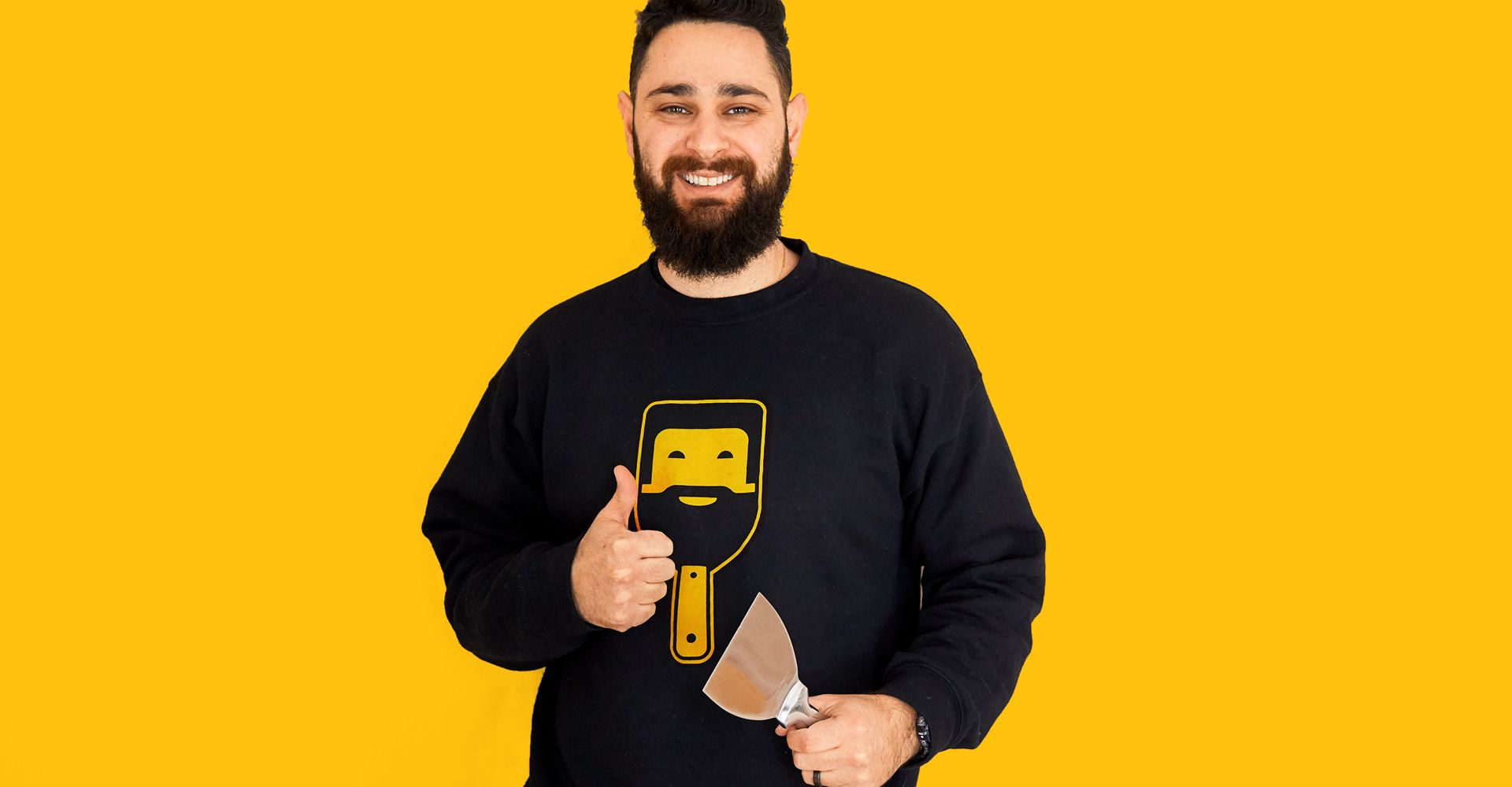 A man with a beard is holding a spatula and giving a thumbs up.