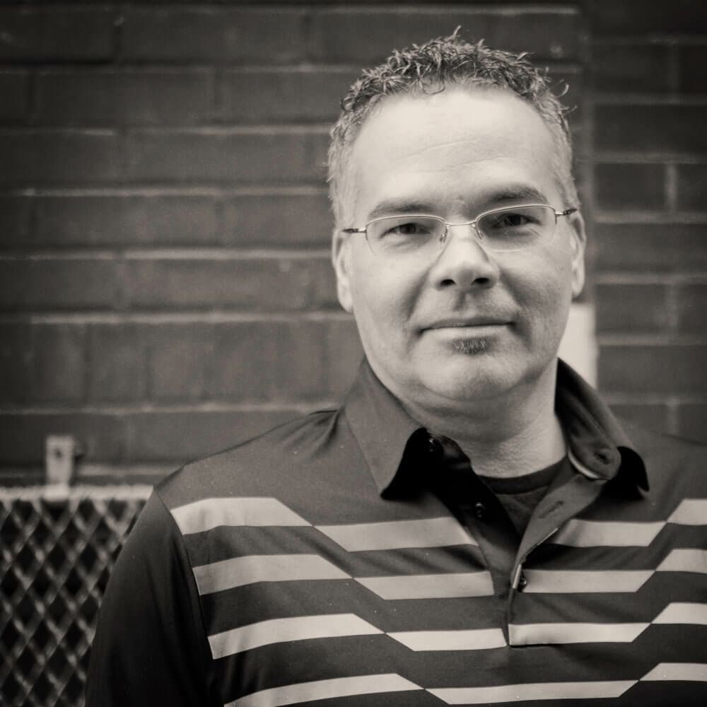 A man wearing glasses and a striped shirt is standing in front of a brick wall