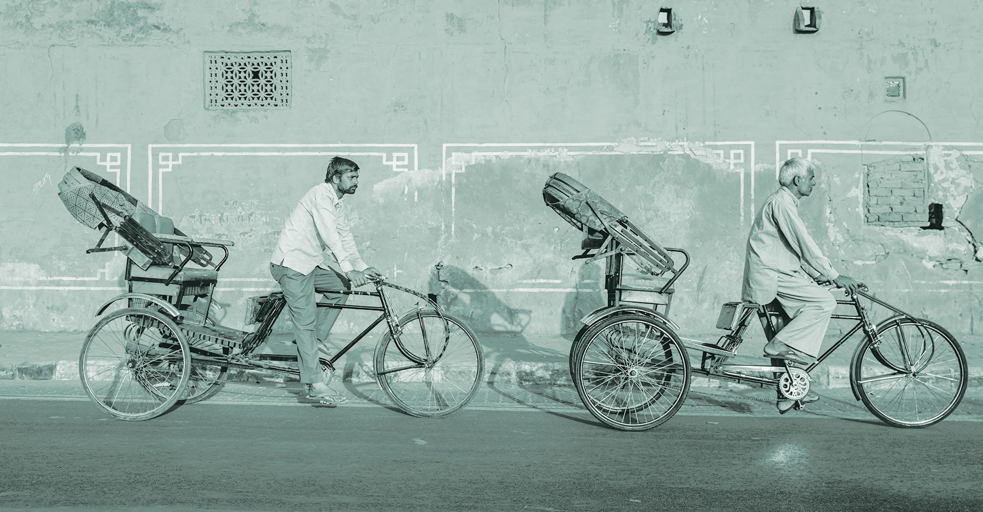 Two men are riding bicycles in front of a wall.