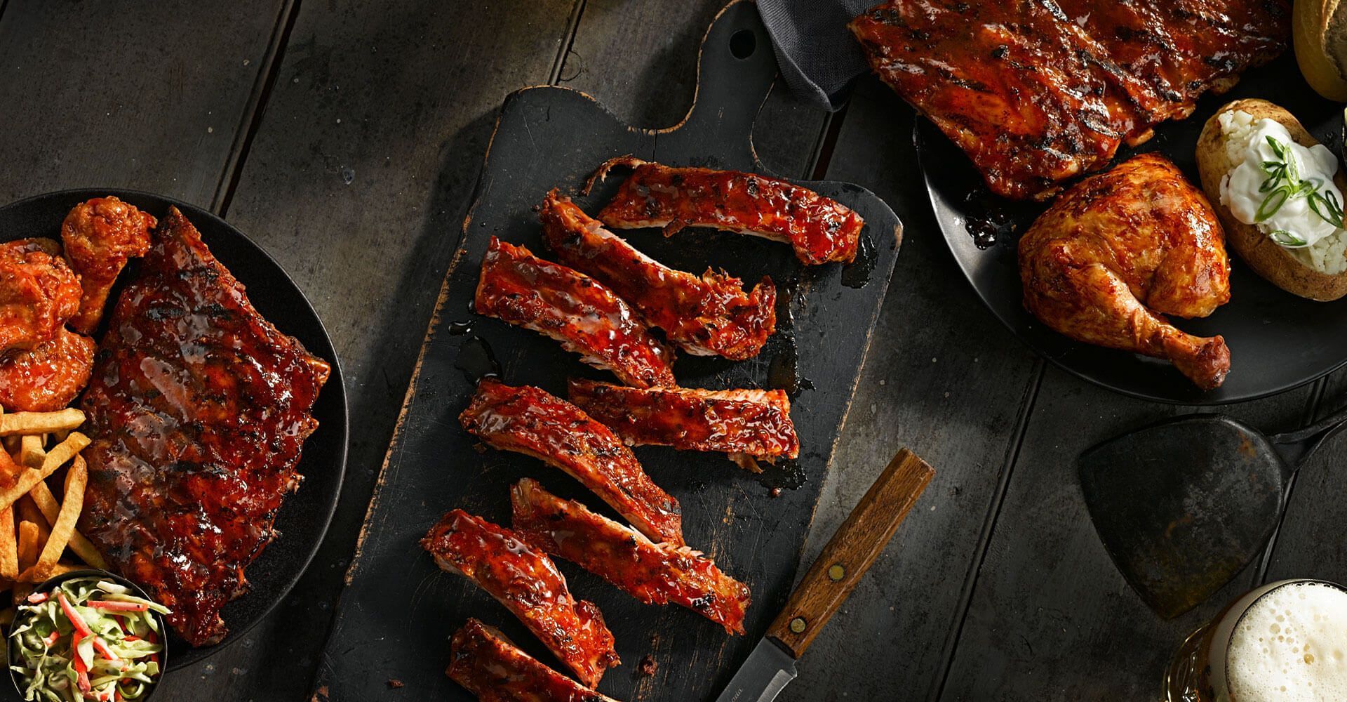 A table topped with plates of food including ribs , chicken , potatoes and coleslaw.