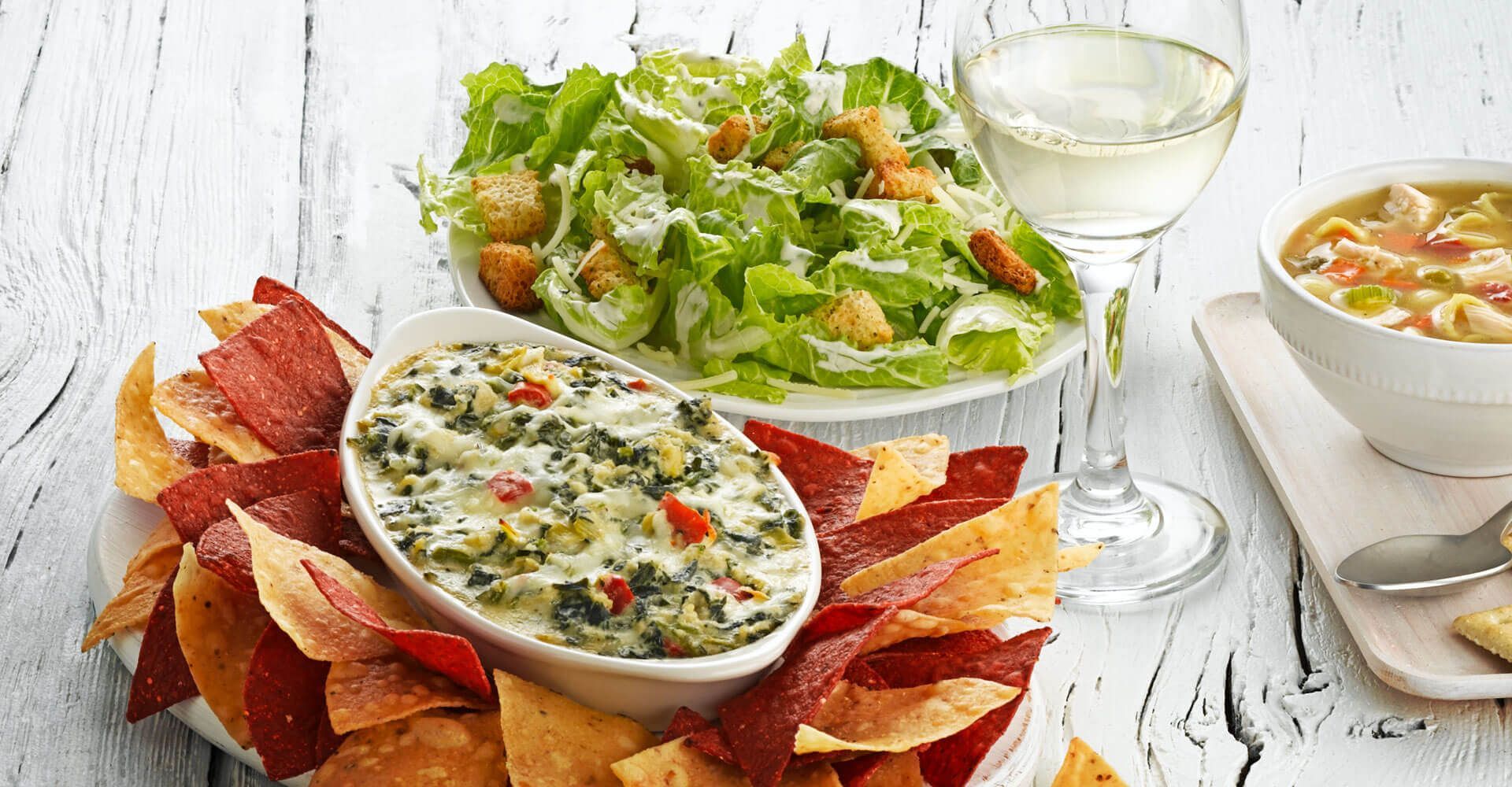 A table topped with a bowl of dip , tortilla chips , a salad and a glass of wine.