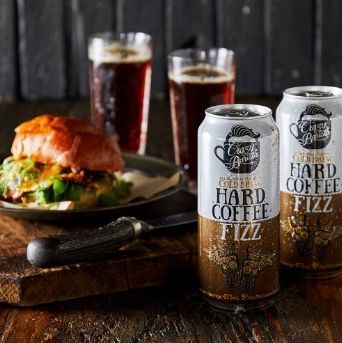 Three cans of hard coffee fizz sit on a wooden table next to a plate of food.