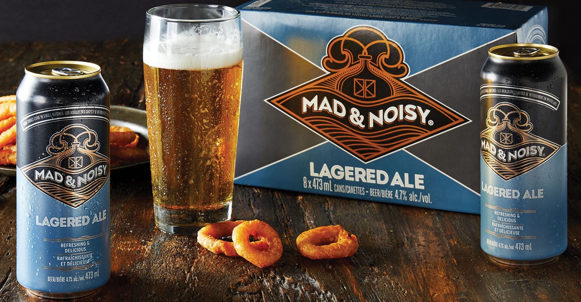 A box of mad & noisy lagered ale next to a glass of beer and onion rings