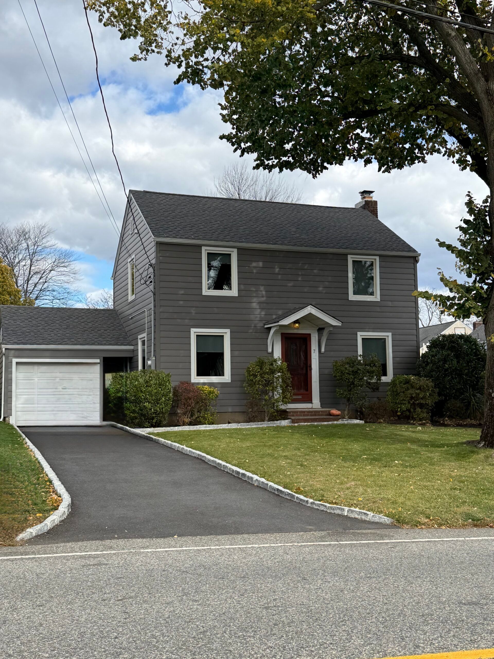 Gray two-story house with a garage and driveway. Green lawn and trees. Cloudy sky.